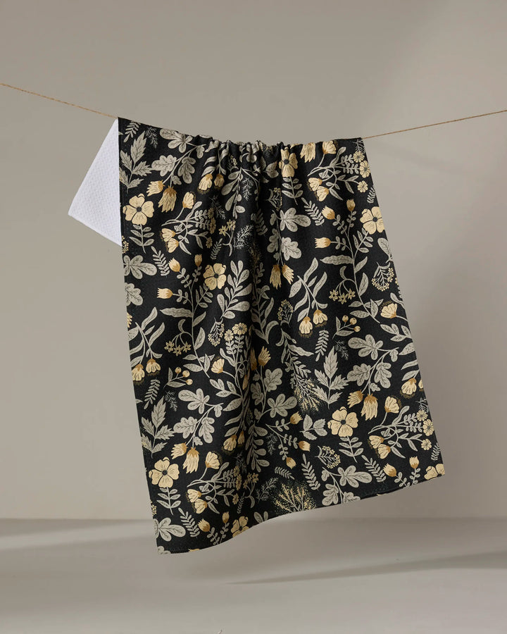 The Danelys Sidron "Woodland Wildflowers" tea towel, featuring a black base with yellow and white floral designs, hangs on a clothesline against a plain backdrop, with a hint of white fabric showing behind.