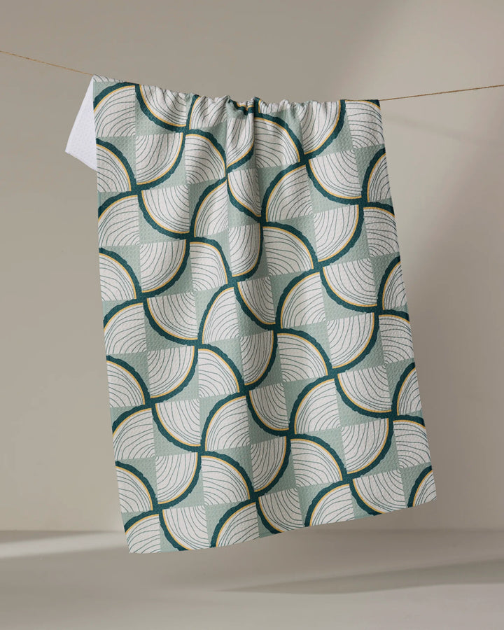Twig + Spark’s “Woodpile” recycled kitchen towel, featuring a green, white, and gold geometric pattern of overlapping curved lines, hangs neatly on a clothesline against a neutral background.