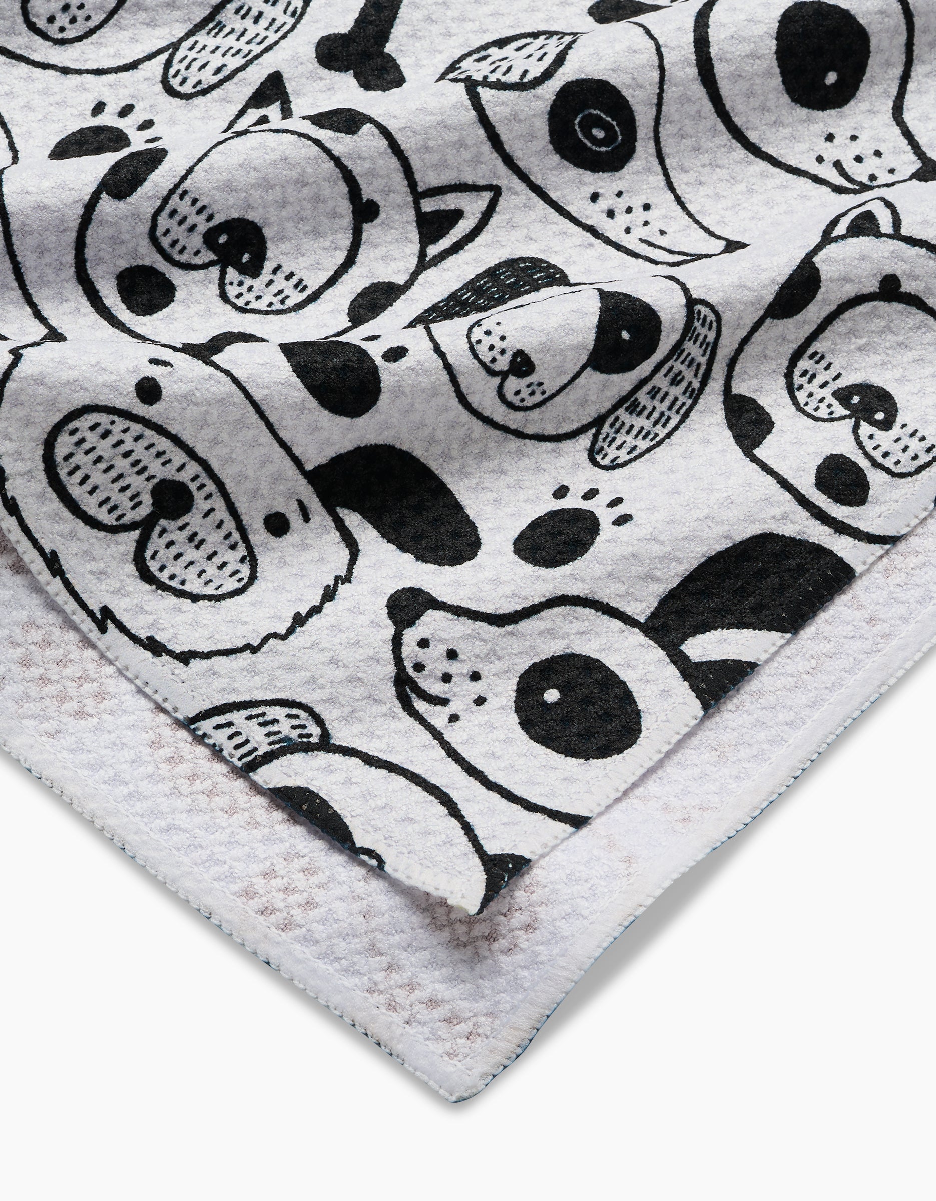A close-up of Jessica Swift’s Woofin Around towel shows ultra-absorbent, textured white Geoweave fabric with a black cartoon animal face pattern—dogs, bears, and more—with overlapping edges in the corner.