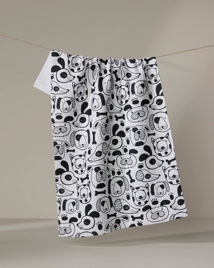 The "Woofin Around" kitchen towel by Jessica Swift features playful black dog faces, bones, and paw prints on white eco-friendly fabric and hangs on a clothesline against a neutral background.