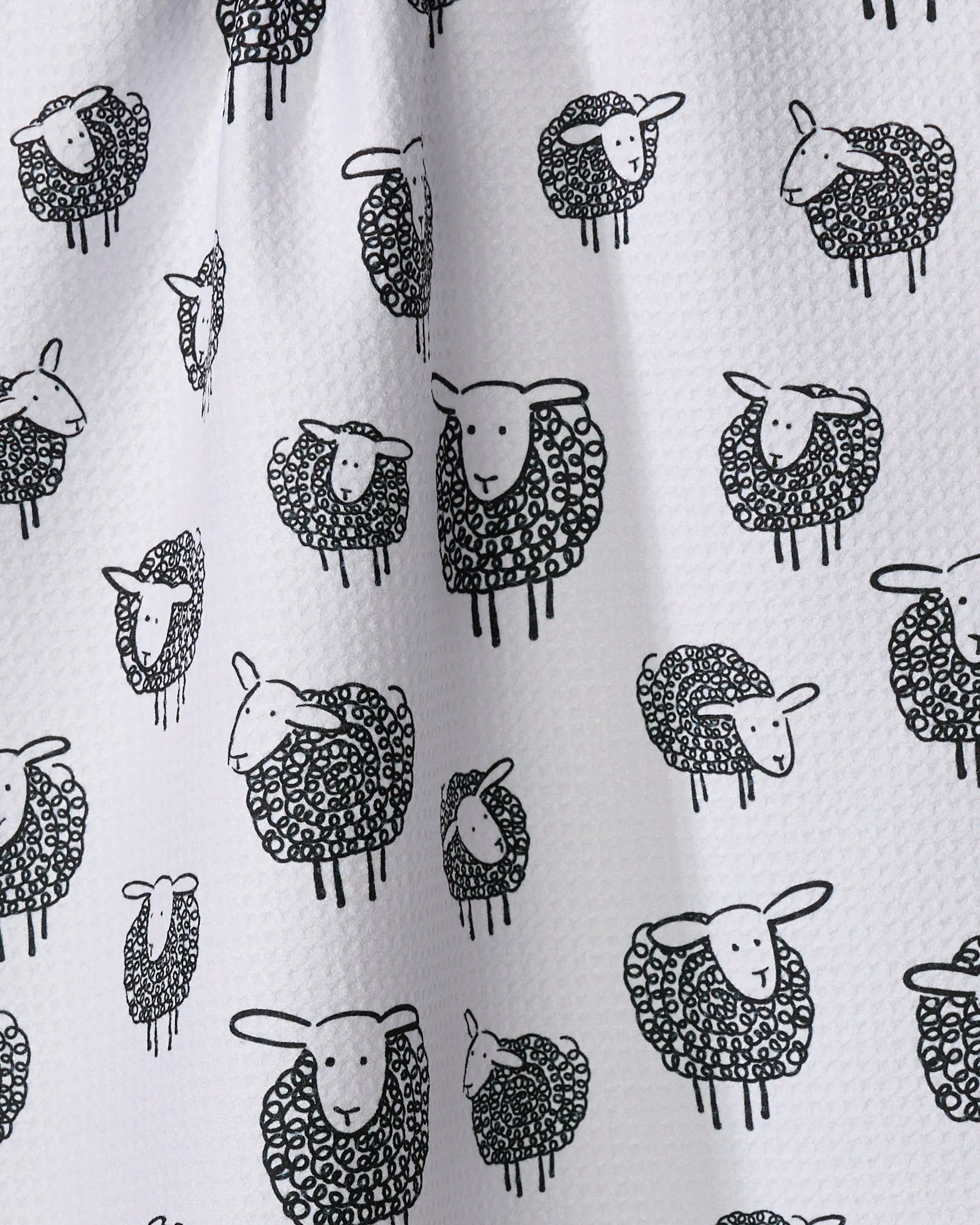 The Kris Ruff Wooly Sheep tea towel features a white textured fabric with a repeating pattern of black-outlined cartoon sheep, each with a curly, shaded body and simple facial features.
