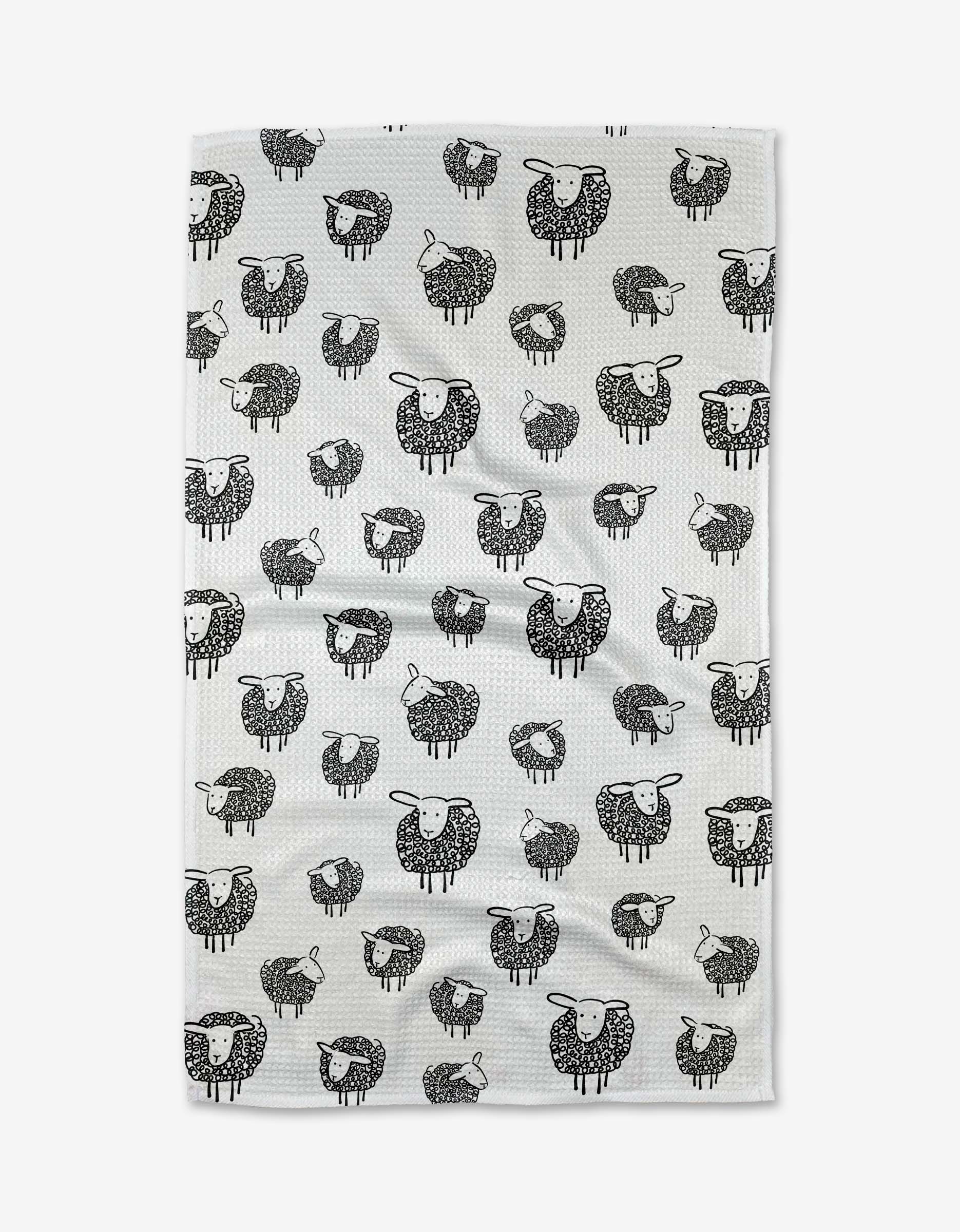 The Wooly Sheep kitchen towel by Kris Ruff is white, made from recycled material, and features an even pattern of black and white cartoon sheep in various poses.