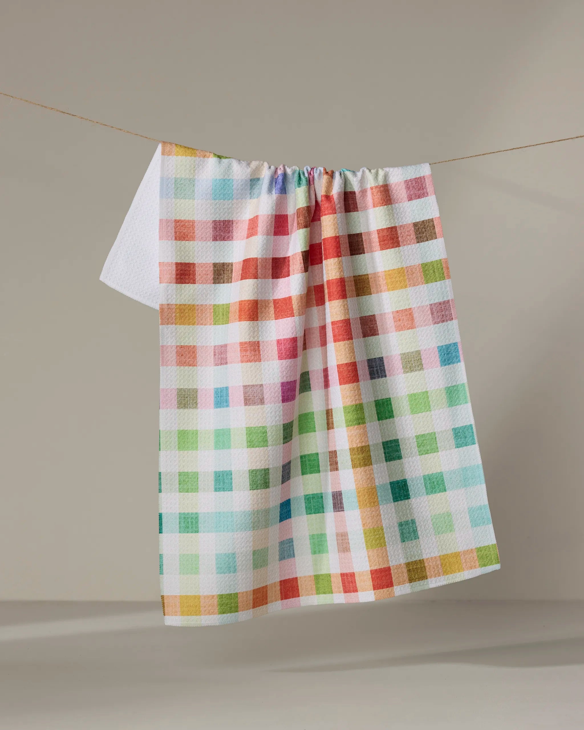 The Pascale Dilger Woven Spring Plaid tea towel features vibrant red, green, blue, yellow, and orange checks—ideal as a quick-drying kitchen towel and shown hanging on a clothesline against a simple beige background.