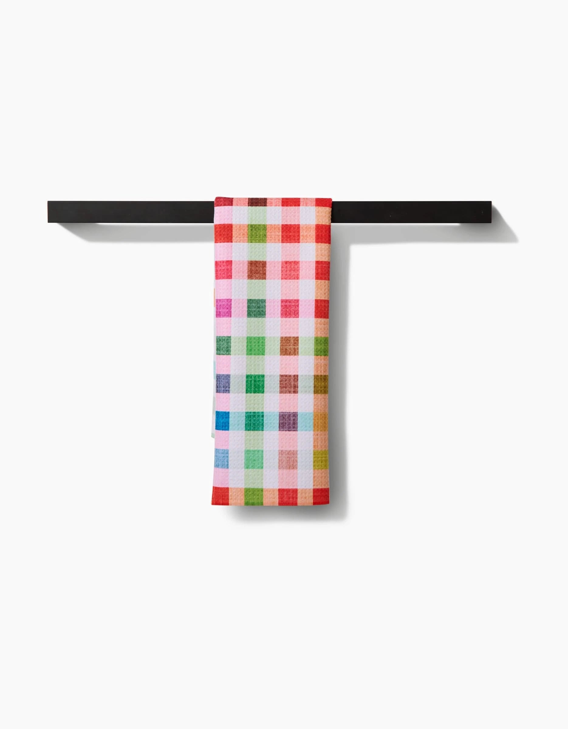The Pascale Dilger Woven Spring Plaid towel, crafted from ultra-absorbent Geoweave fabric and featuring vibrant red, pink, green, yellow, and blue squares, hangs neatly from a simple black bar against a white background.
