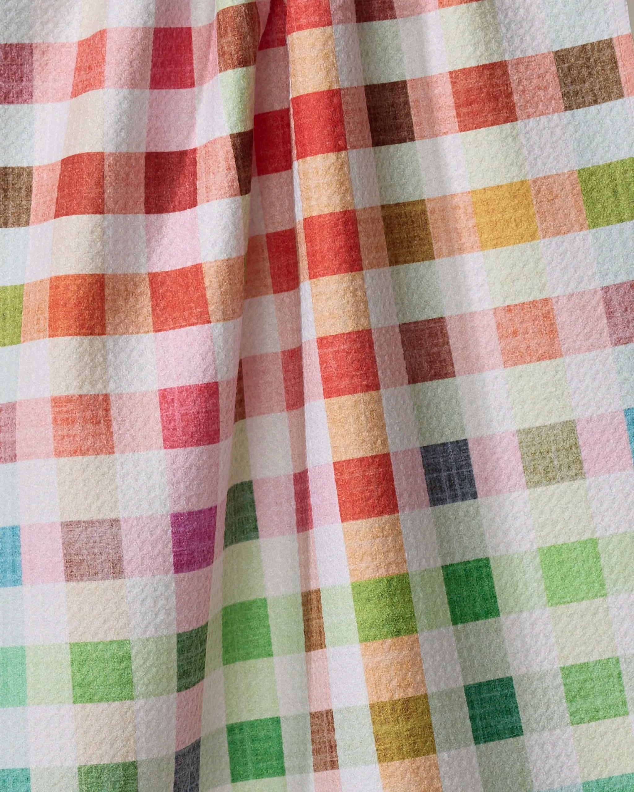 A close-up of Pascale Dilger’s Woven Spring Plaid recycled kitchen towel shows a textured surface with a vibrant checkered pattern in red, orange, yellow, green, pink, and gray squares on white.