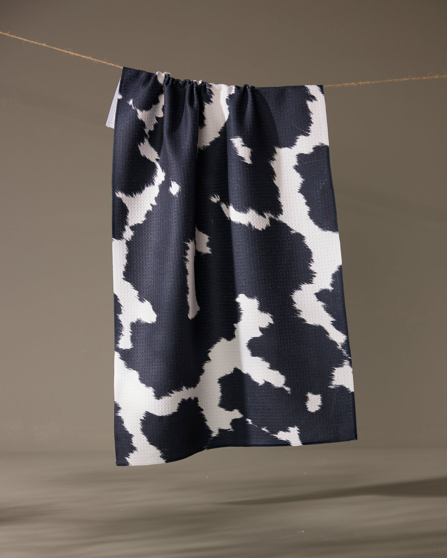 The Geometry Wrangler tea towel by Geometry, featuring a black and white cow print and made from recycled materials, hangs on a string against a neutral background.