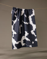 The Geometry Wrangler tea towel by Geometry, featuring a black and white cow print and made from recycled materials, hangs on a string against a neutral background.