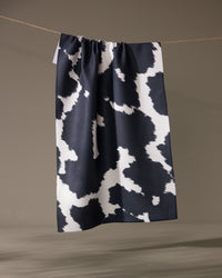 The Geometry Wrangler tea towel by Geometry, featuring a black and white cow print and made from recycled materials, hangs on a string against a neutral background.