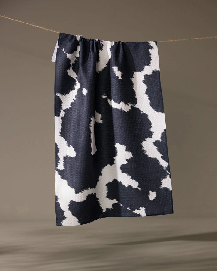 The Geometry Wrangler tea towel by Geometry, featuring a black and white cow print and made from recycled materials, hangs on a string against a neutral background.