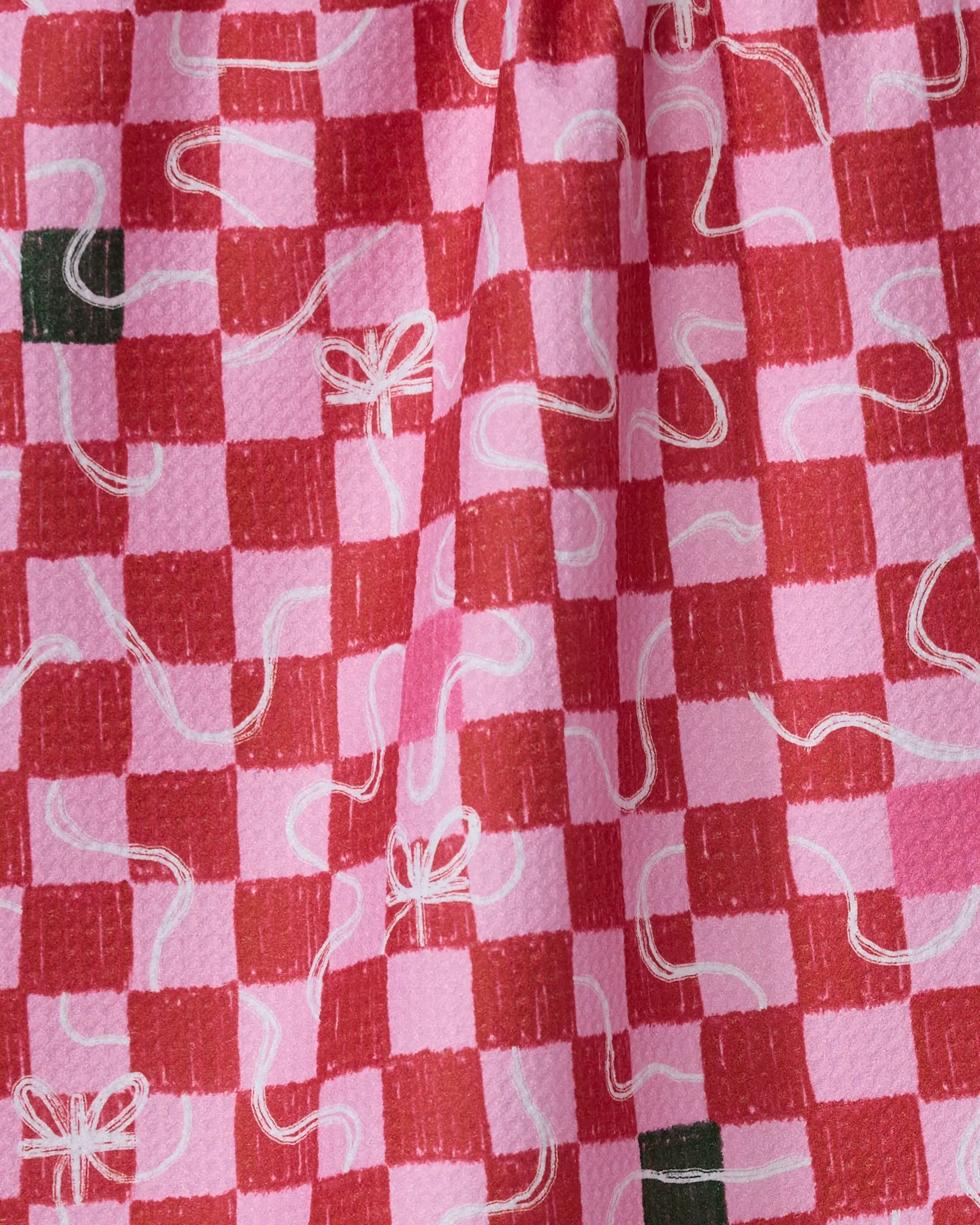 A close-up of Geometry's Wrapped Checkers tea towel shows pink and red checks, green squares, and white ribbon accents on an ultra-absorbent, textured fabric.
