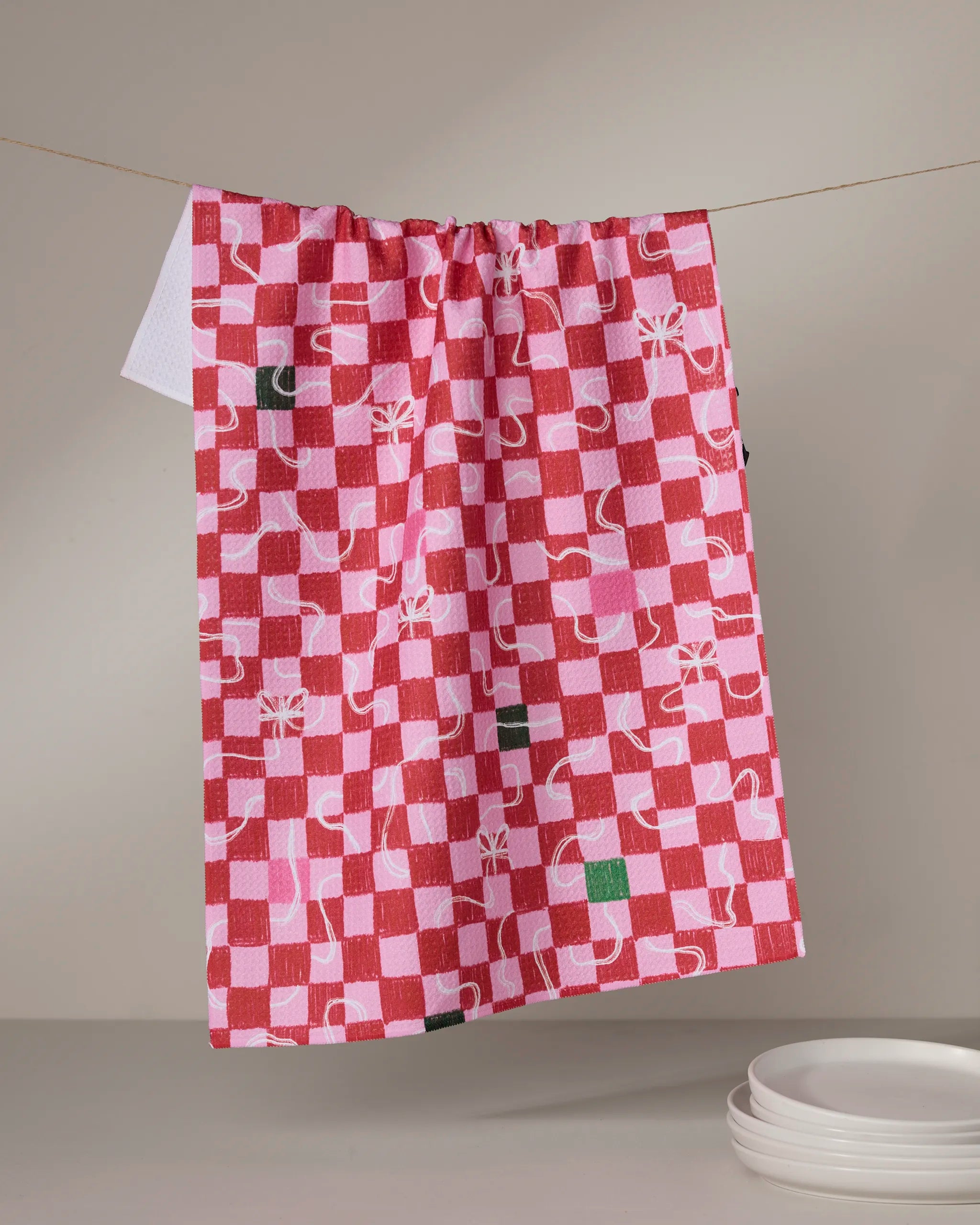 The Geometry Wrapped Checkers tea towel, featuring red and pink abstract checkered patterns and made from recycled materials, hangs above white plates against a neutral background.