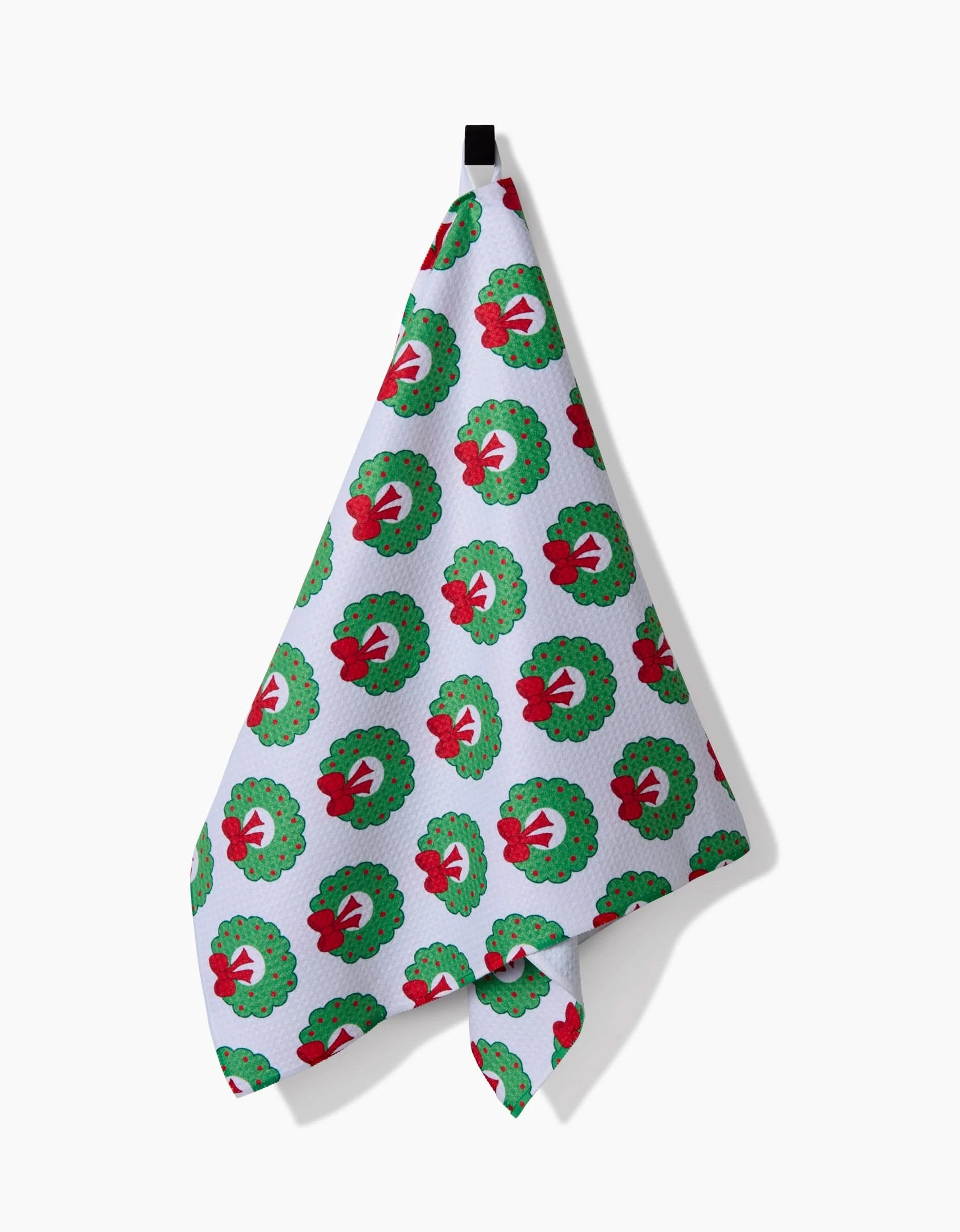 The Mindfulnice Wreath Wishes kitchen towel, crafted from recycled materials and adorned with a green wreath and red bow pattern, hangs from a black hook against a white background.