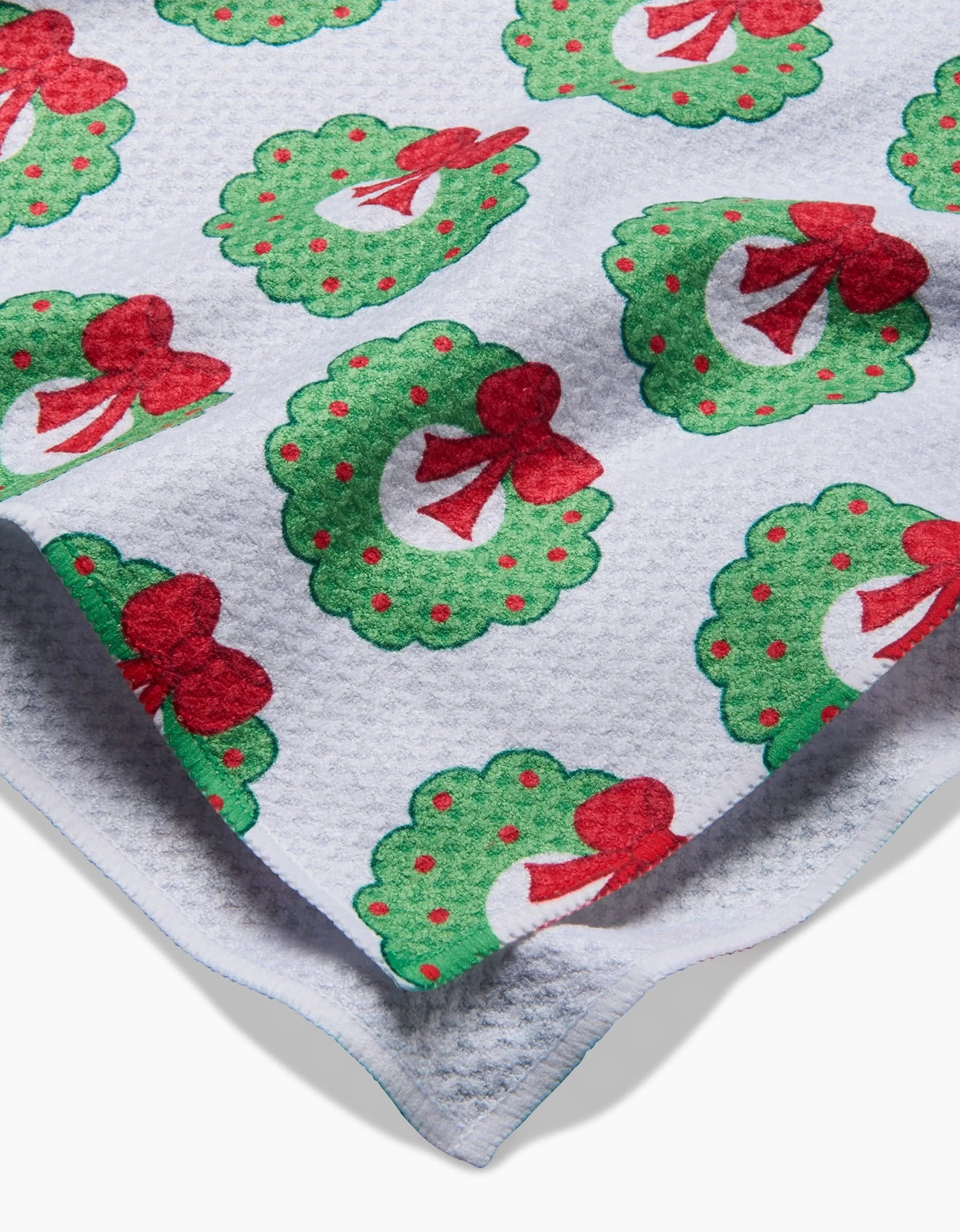 The Mindfulnice Wreath Wishes tea towel features a white textured fabric printed with green Christmas wreaths adorned with red bows and berries. Slight folding at the corner highlights its festive charm and quality.