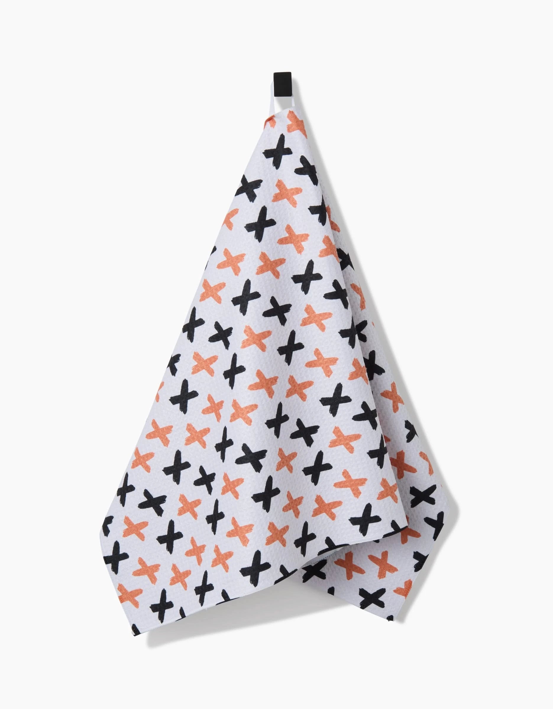 The X Marks The Spot Orange tea towel by Charlie Rowan Designs, featuring a white background with scattered black and orange X marks, hangs from a black loop against a plain backdrop.