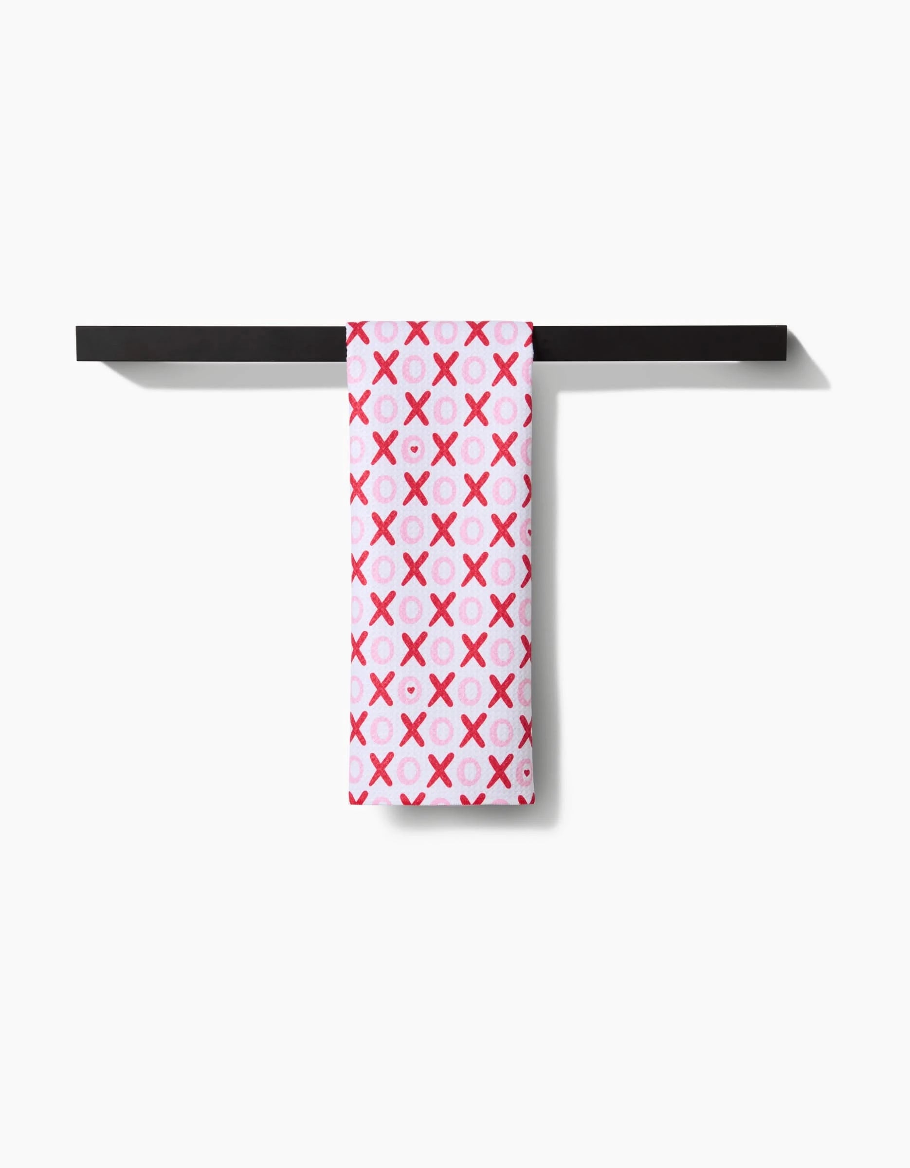 The XOXO tea towel by Sarah Daugherty, featuring a pink and red X and O pattern, hangs neatly over a black bar against a white background.