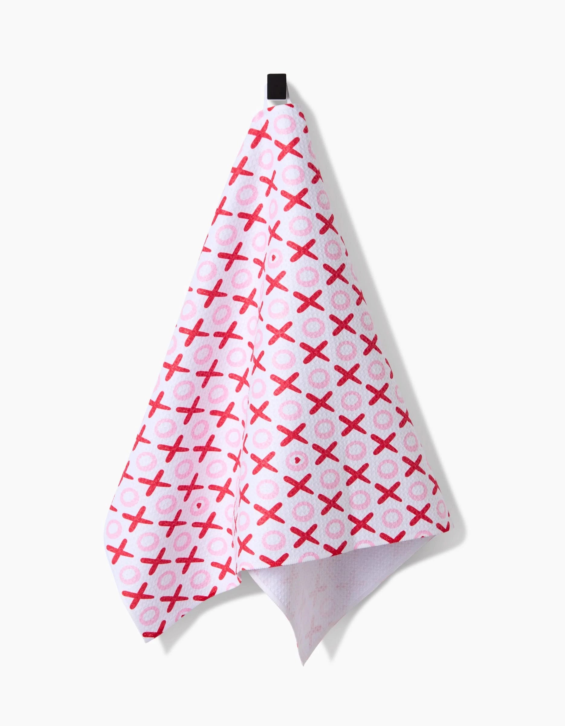 The XOXO tea towel by Sarah Daugherty hangs from a black hook, featuring a pink and red XO pattern. Made from quick-drying recycled materials, it’s both eco-friendly and charming.