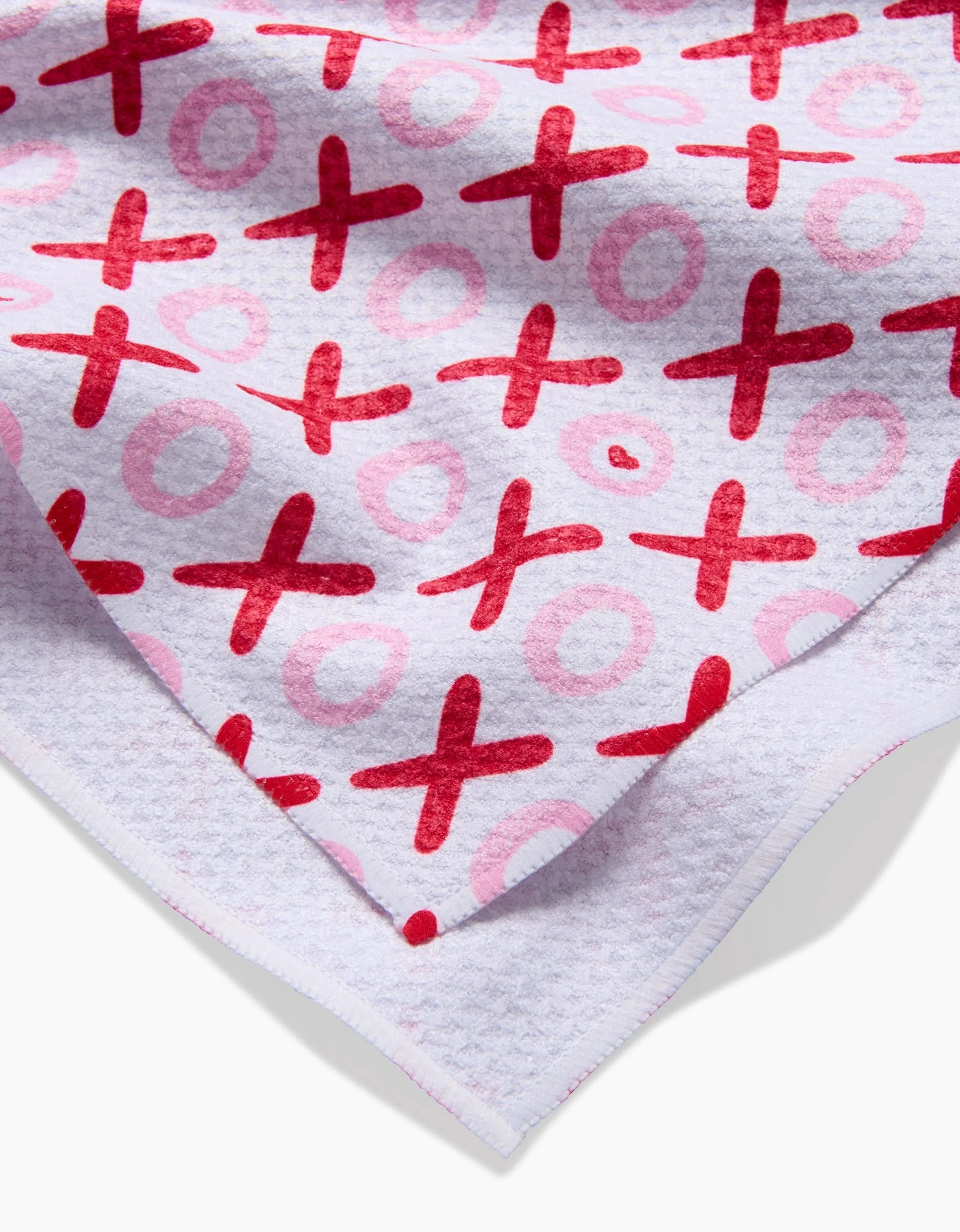 A close-up of the XOXO tea towel by Sarah Daugherty shows its red and pink Xs and Os pattern on a light, textured background, with a folded corner displaying the reverse side.