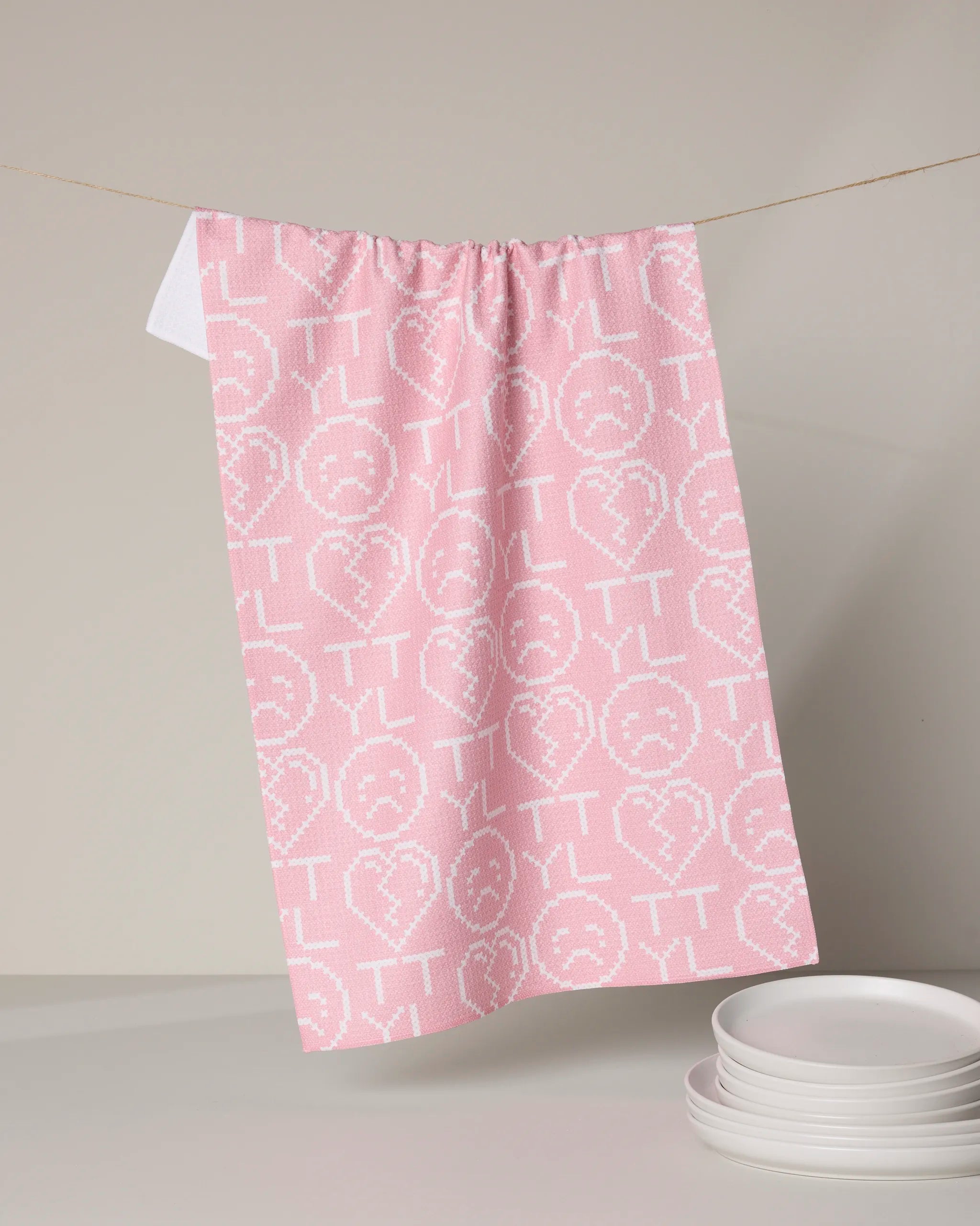 The XOXOX Emoticon tea towel by Geometry, made from recycled materials, showcases TTYL and sad face icons in pink and hangs above white plates against a neutral background.