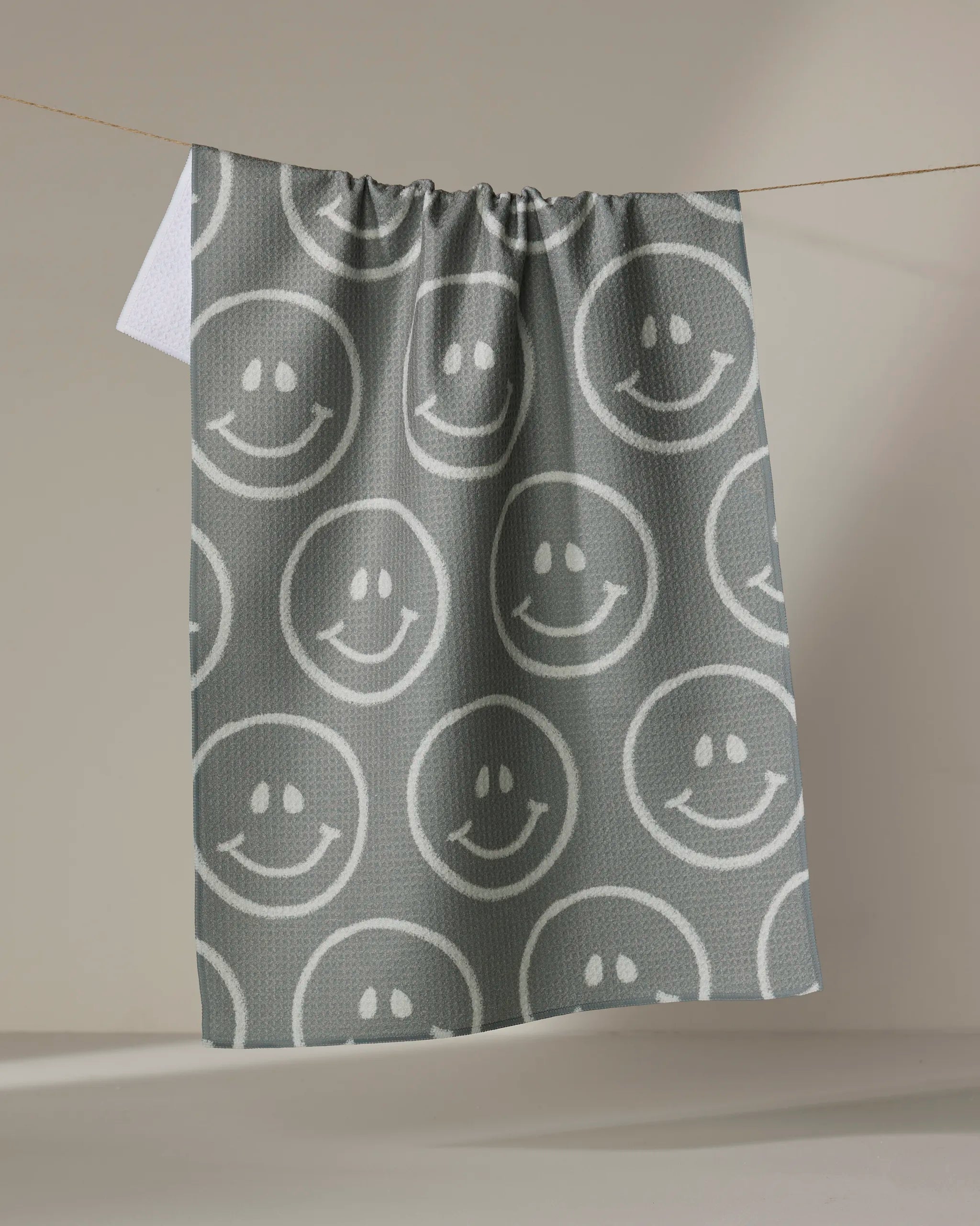 The "You Make Me Happy" towel by Kelsi Fullmer, featuring a gray ultra-absorbent fabric with white smiley faces, hangs on a clothesline against a plain beige background.