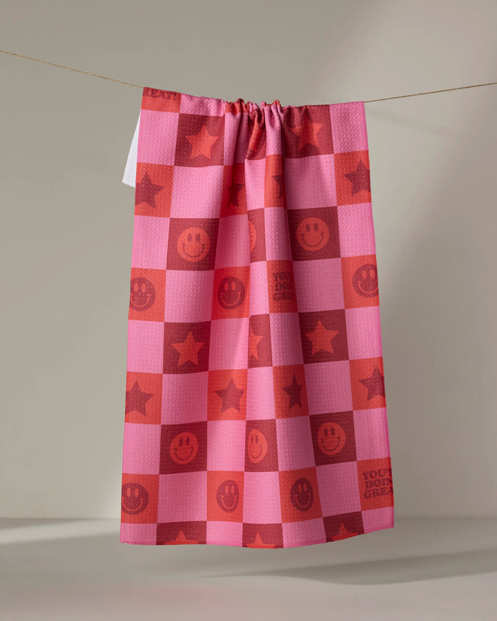 The "You're Doing Great" tea towel by Quotes by Christie features pink and red checkers, smiley faces, stars, and uplifting text, hanging against a beige background. It's crafted from recycled materials.