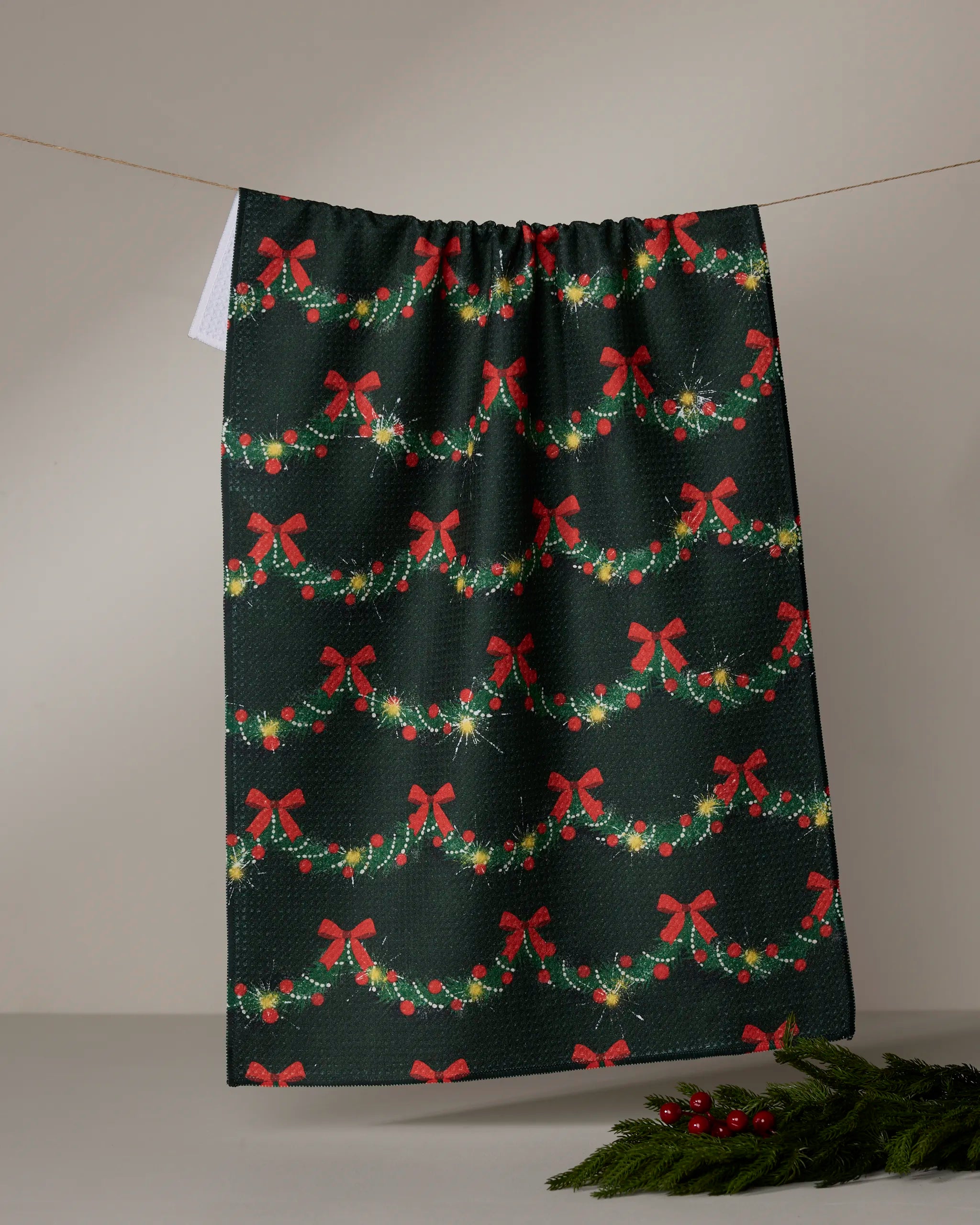 The Geometry Yuletide Garland tea towel in dark green hangs on a line, featuring red bows, lighted garlands, and holly. Greenery and red berries sit nearby for a festive look.