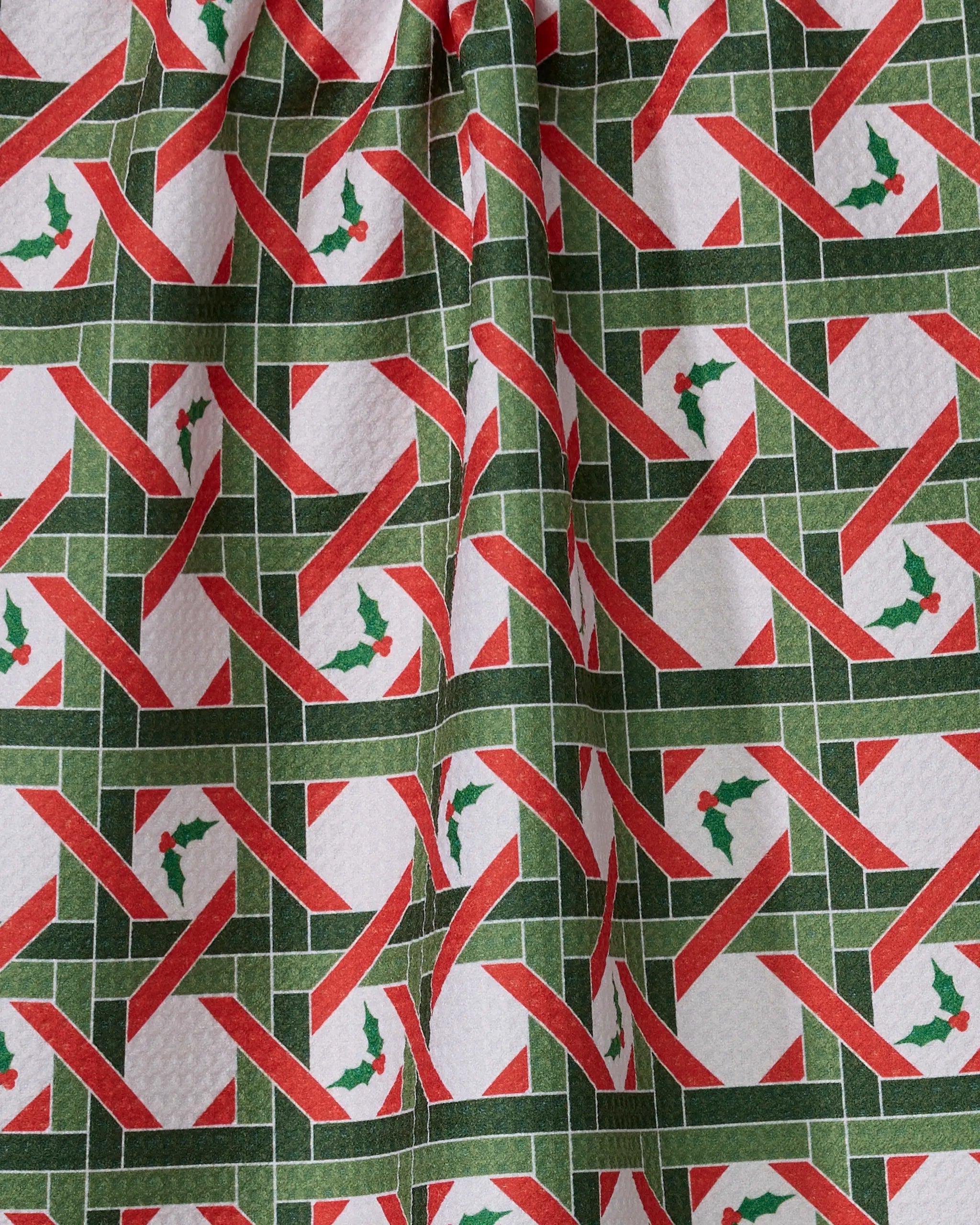 The Geometry Yuletide Lattice kitchen towel is made from recycled materials and features intersecting green and red stripes with holly leaf and berry motifs on white, shown draped with visible folds.