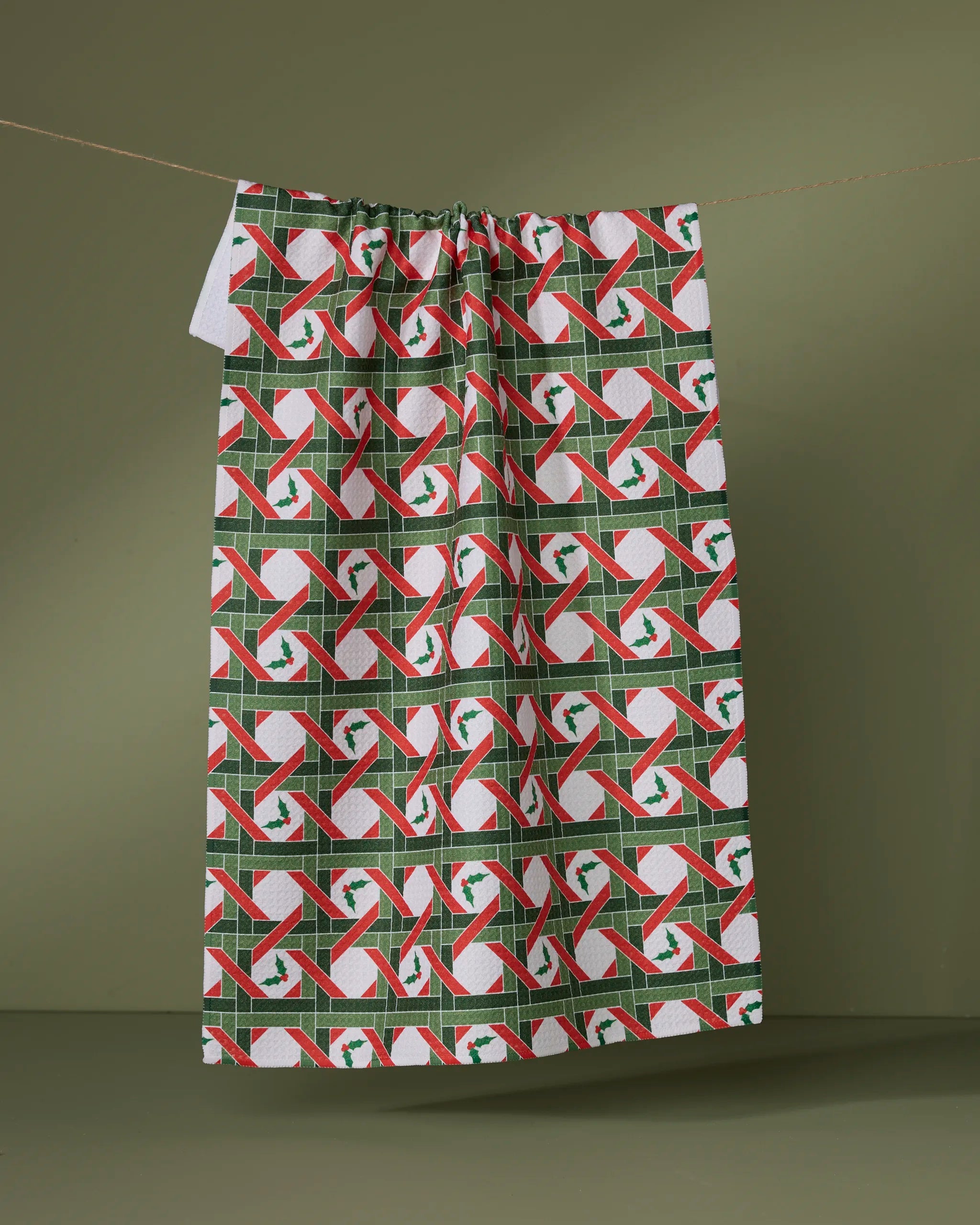 The Yuletide Lattice kitchen towel by Geometry, made from recycled materials, displays candy canes, holly wreaths, and red, white, and green geometric patterns on an olive green background.