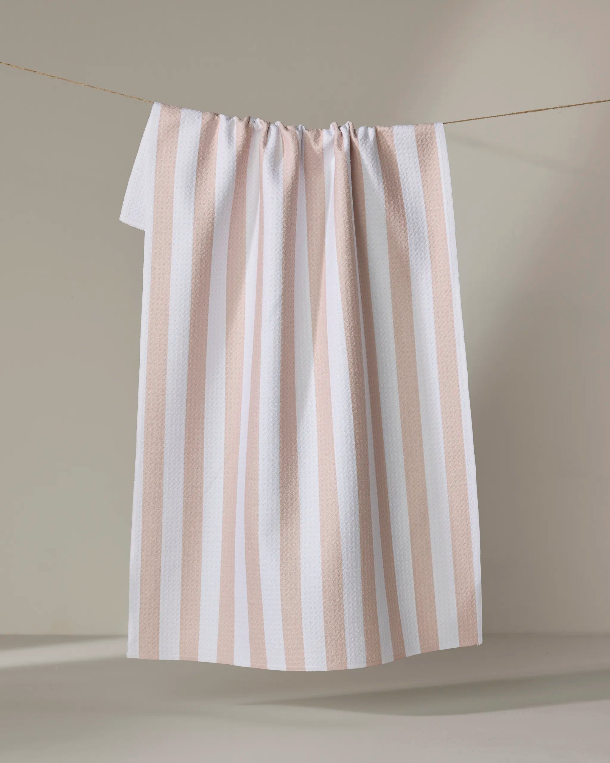The Yvonne Stripe kitchen towel by Kailee Wright, featuring white and light beige stripes and made from recycled materials, hangs from a thin rope against a plain, light backdrop.