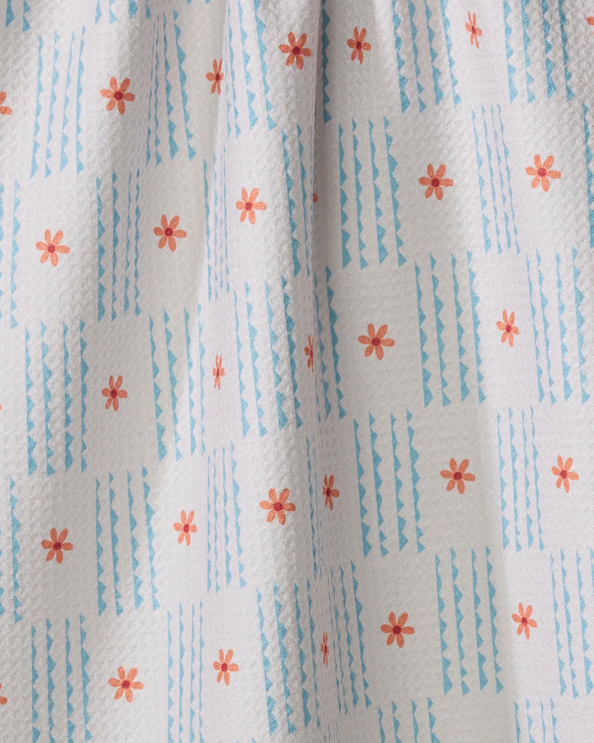 A close-up of the Ziggy Floral tea towel by Elizabeth Edwards, featuring small orange flowers and blue geometric stripes in a grid on white. Its slightly dimpled texture highlights its ultra-absorbent quality.