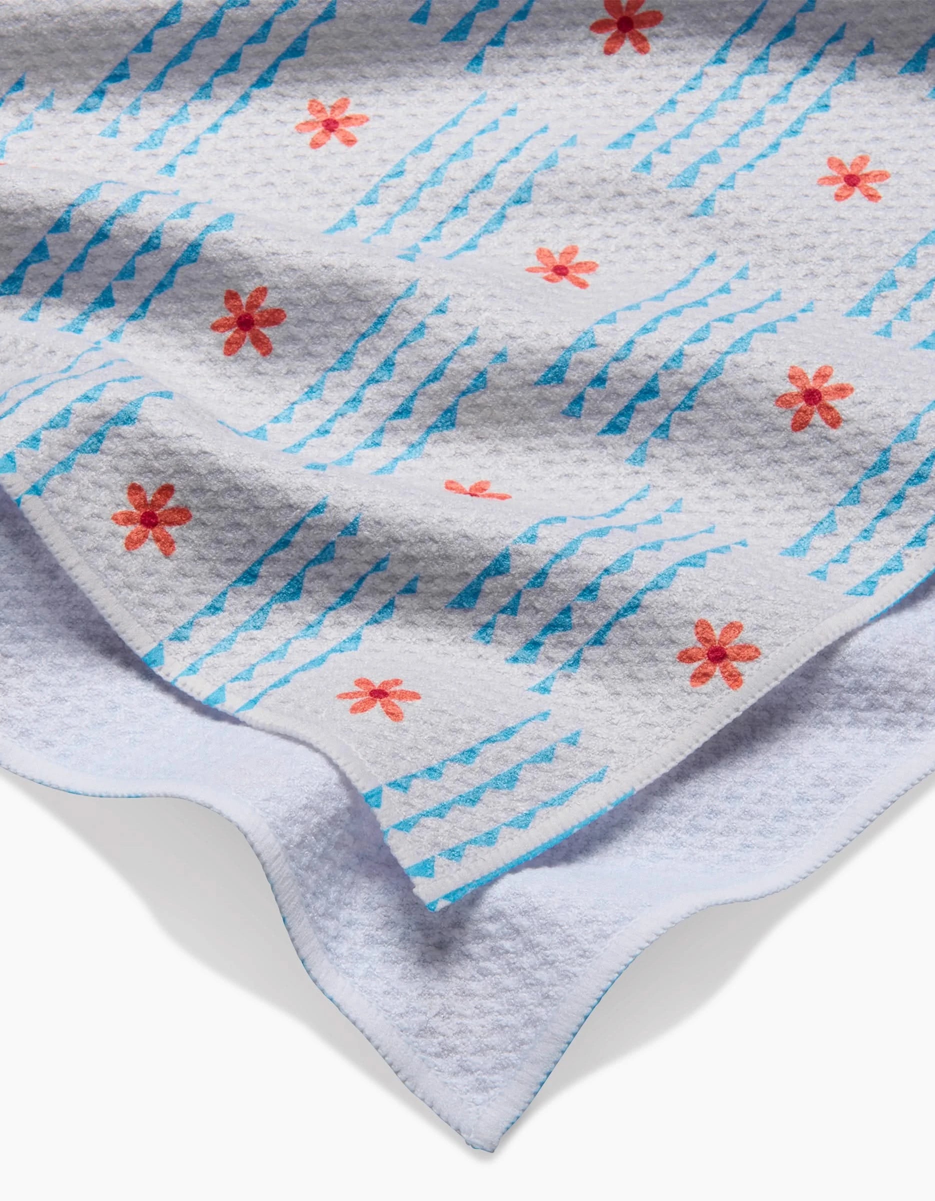 A close-up of the Ziggy Floral tea towel by Elizabeth Edwards, featuring red-orange flowers and blue geometric lines on textured Geoweave fabric, with its edge folded to show the plain white underside.