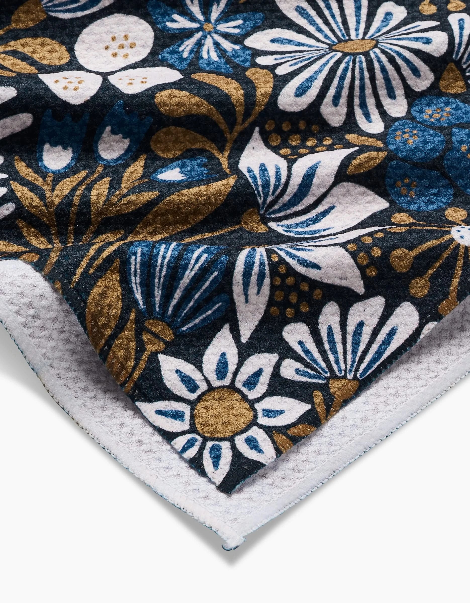 A close-up of the Zillia tea towel by Amy MacCready, featuring a floral pattern in white, blue, and gold on a dark background. The Geoweave fabric’s textured edge is folded over, exposing the white underside.