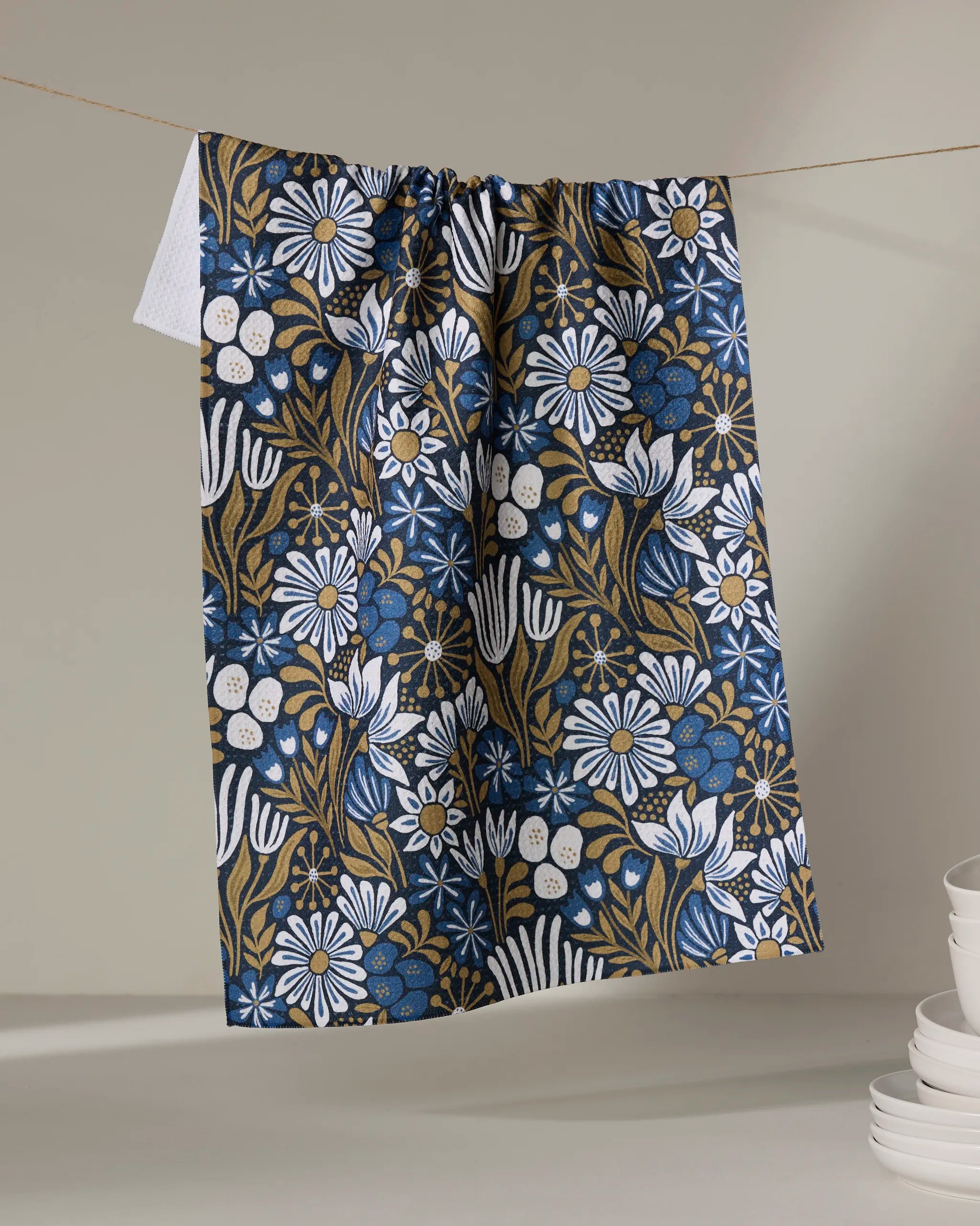 The Amy MacCready Zillia tea towel, ultra-absorbent with a botanical print in blue, white, and mustard yellow, hangs on a line; white plates are stacked in the lower right corner.