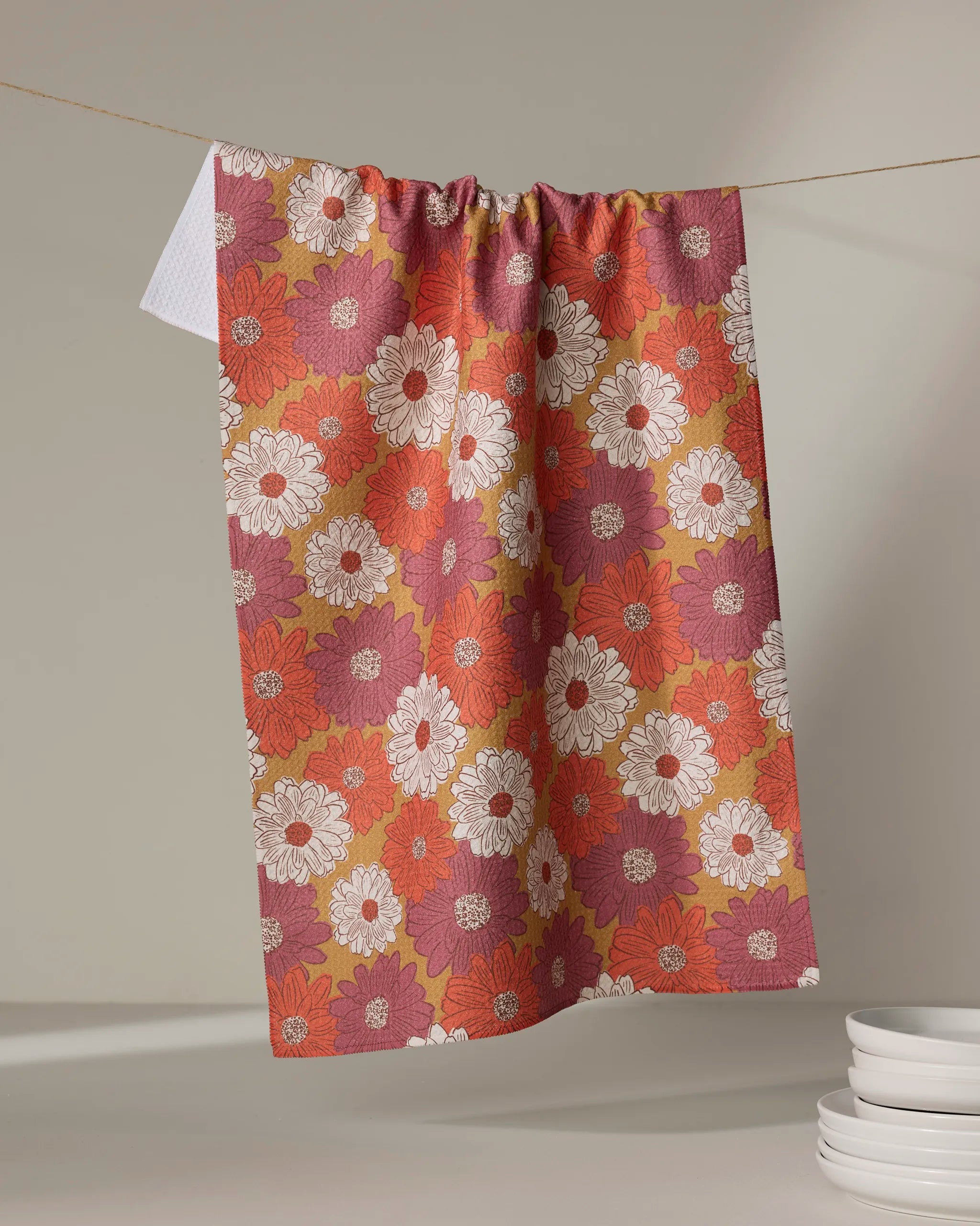 The Kayla Ann Zinnia Forever dish towel, featuring a floral pattern in red, orange, white, and yellow, hangs on a line with stacked white plates in front against a neutral background.
