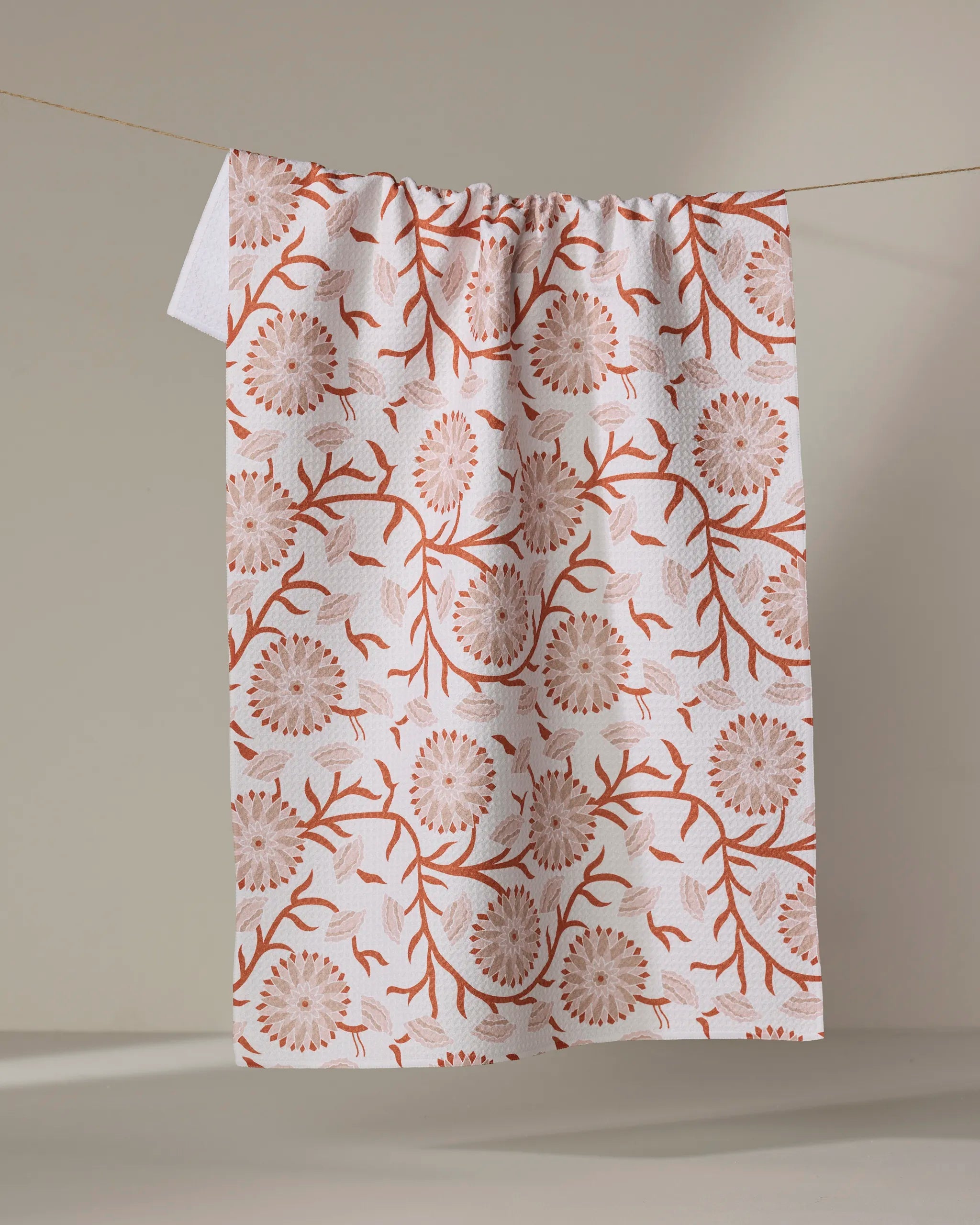 The Zinnie tea towel by Dabney Lee, featuring an orange floral and branch pattern on white Geoweave fabric, hangs on a clothesline against a neutral background.