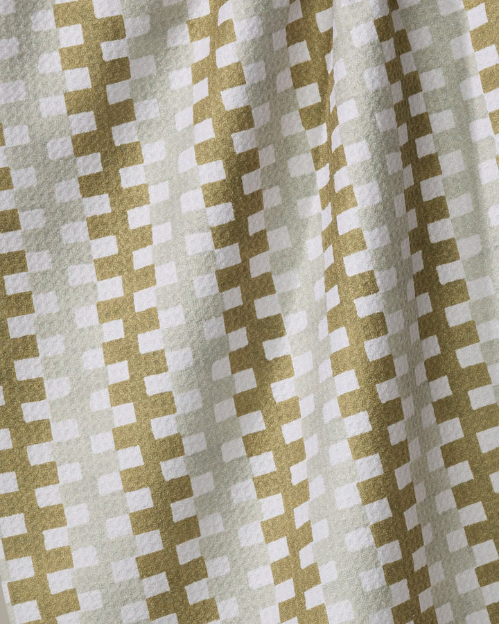 A close-up of Zippy by Studio Jeannot shows its quick-drying recycled fabric, featuring a geometric pattern of white rectangles on light green. Gentle folds and subtle shadows enhance the textured, repetitive design.