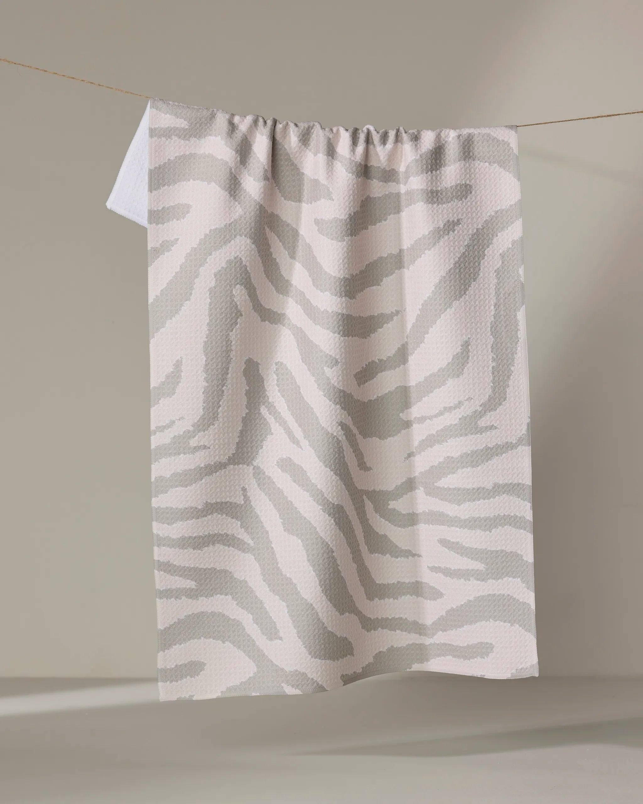 The Zira Gray tea towel by Geometry features a beige and light gray zebra stripe pattern, made from recycled materials, and is displayed hanging on a string against a neutral background.