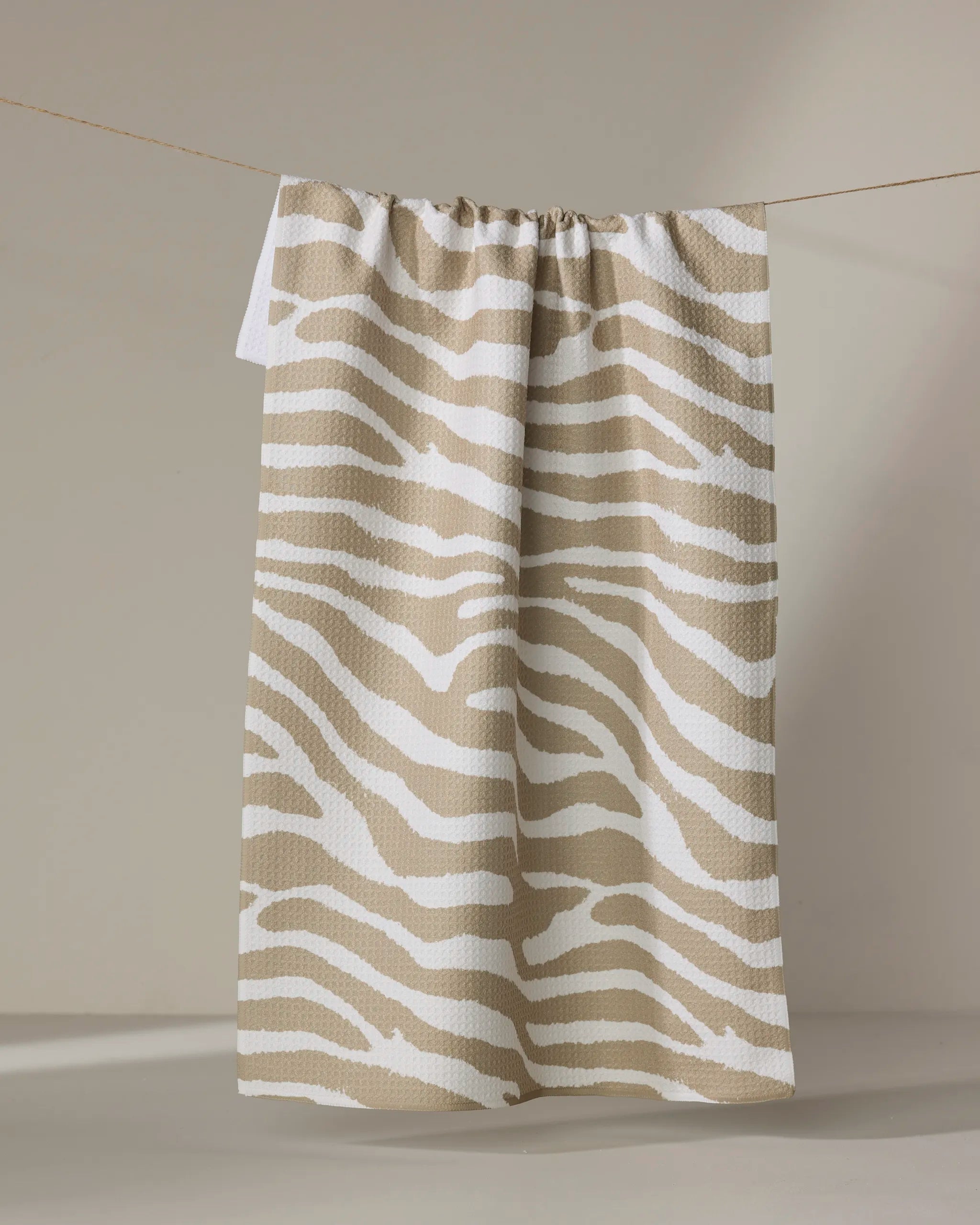The Geometry Zira Tan tea towel, featuring beige and white zebra stripes, hangs on a thin clothesline against a softly lit neutral background.