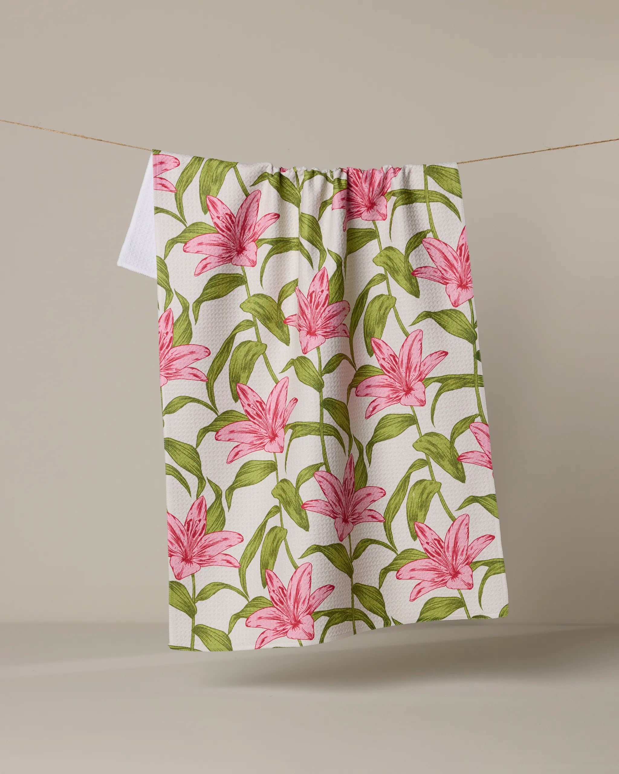 The Geometry Mother Lily kitchen towel, featuring a pink lily and green leaves design and made from recycled materials, hangs on a clothesline against a plain beige background.