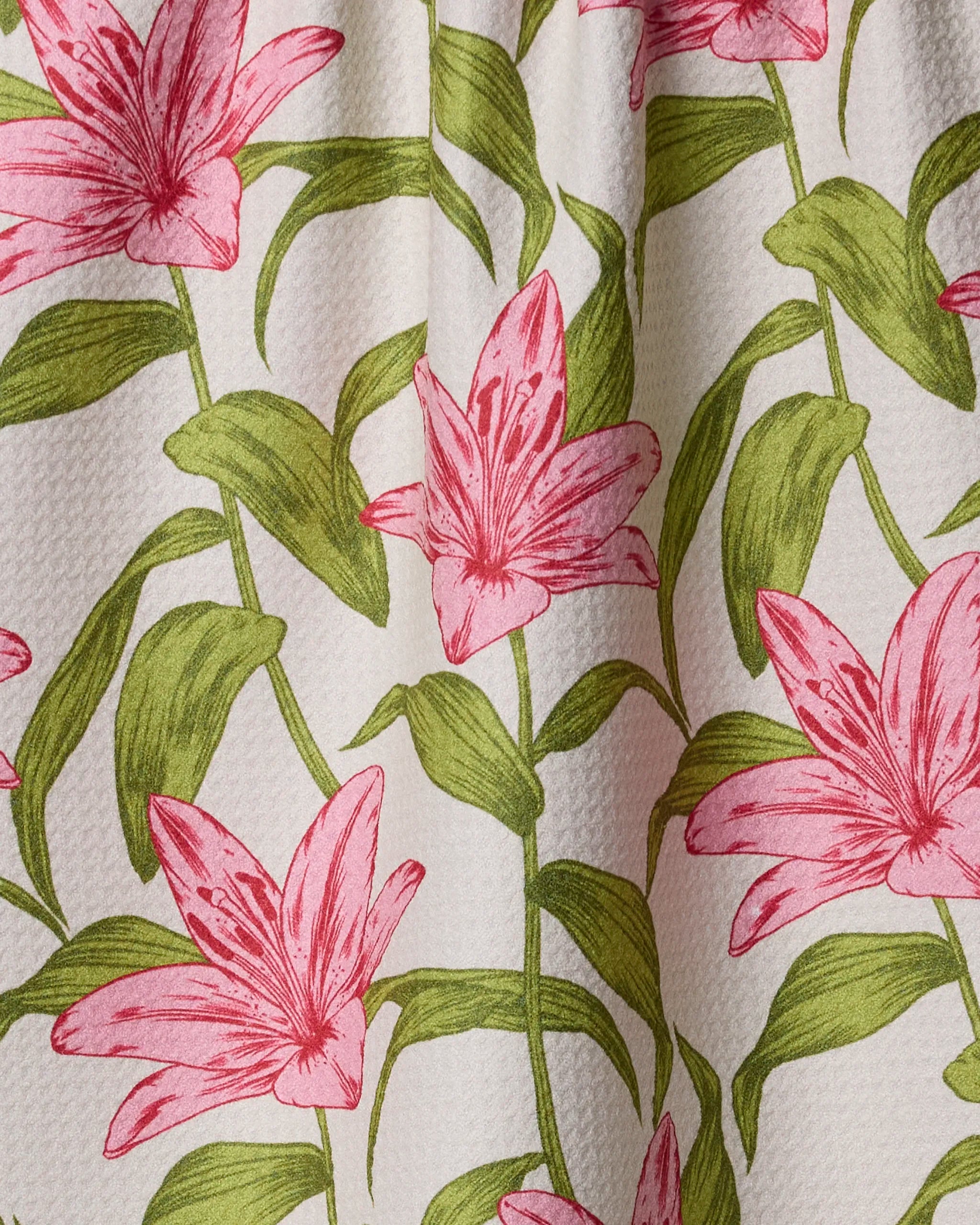 The Geometry “Mother Lily” kitchen towel features pink lilies and green leaves in a detailed, natural style on recycled materials, creating a vibrant botanical design on a white background.