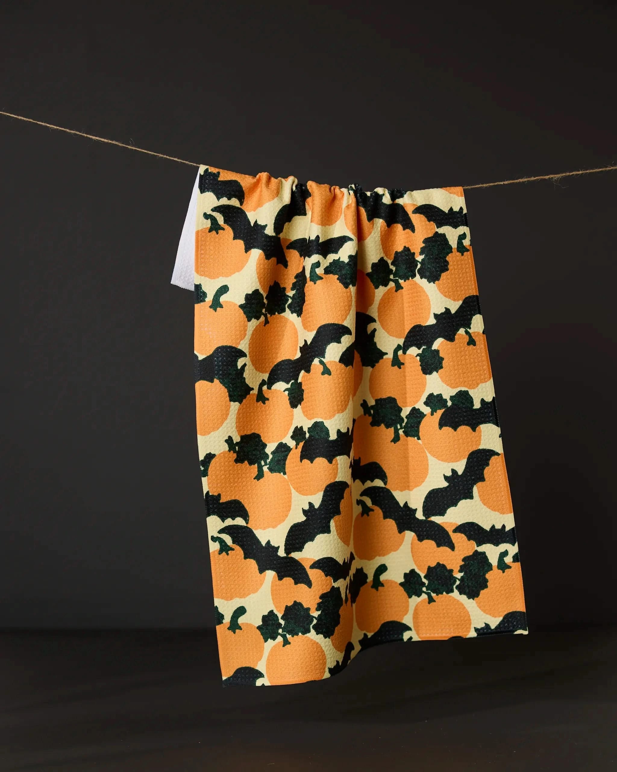 The Geometry “Bat Flight” tea towel, made from recycled materials and adorned with orange pumpkins and black bats, hangs against a dark background.