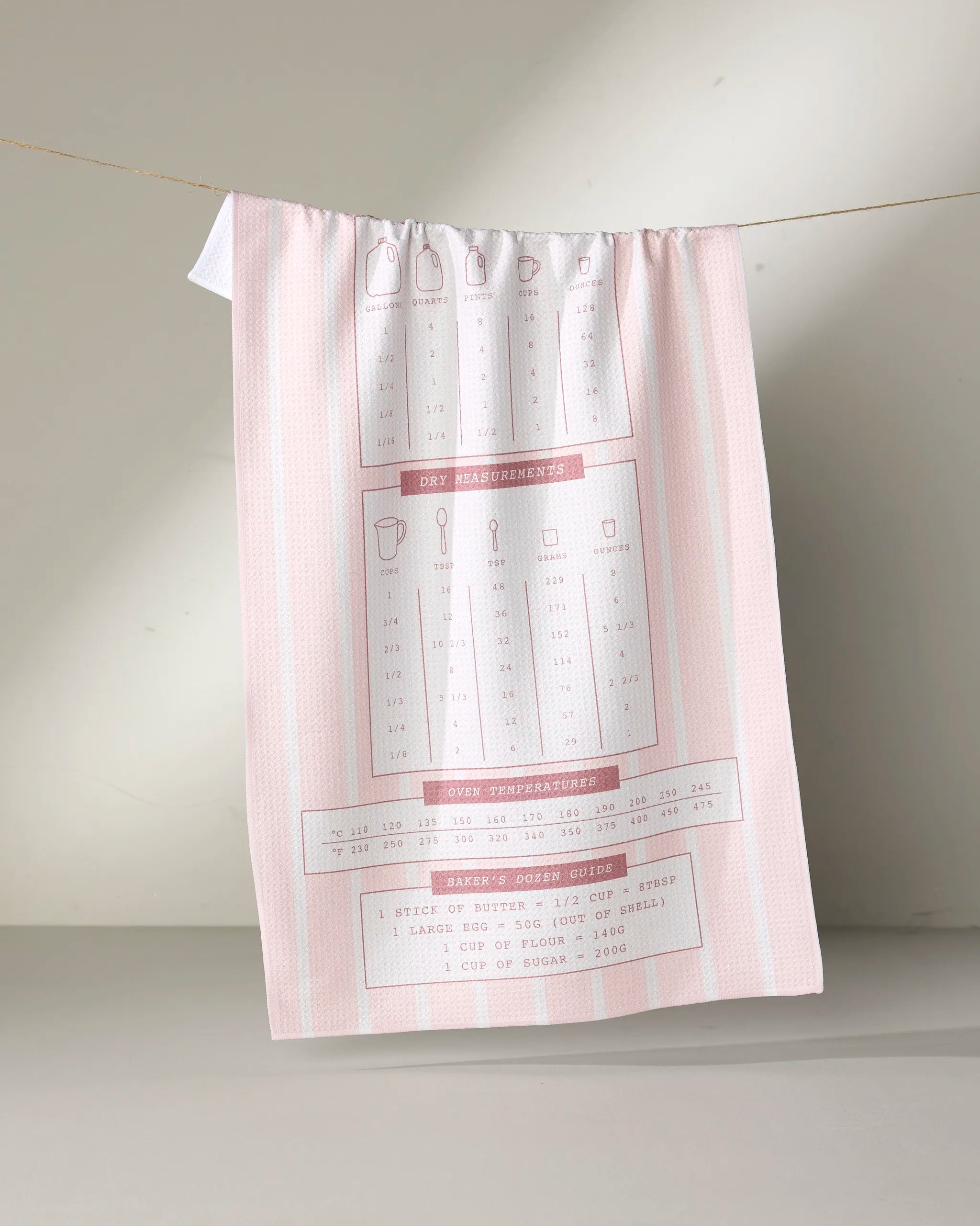 The Brooke Homec Cheat Sheet tea towel, crafted from recycled materials in pink and white, hangs from a line and features red and gray baking measurements plus oven conversions. Ultra-absorbent, it doubles as a handy kitchen reference guide.