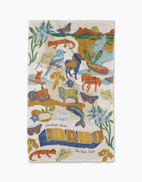 The Greetings From Idaho tea towel by Geometry is crafted from durable geoweave fabric with recycled materials and features colorful Idaho wildlife, flowers, mountains, rivers, and Greetings from IDAHO The Gem State in bold at the bottom.