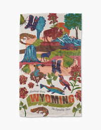 The Geometry Greetings From Wyoming tea towel, made with ultra-absorbent Geoweave™, features illustrations of Wyoming symbols—bison, mountains, cowboy, wildlife, flowers, and trees—with text: Greetings from WYOMING, The Equality State.