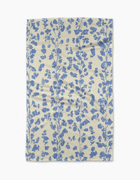 The Lilou Cream kitchen towel by Geometry is eco-friendly and cream-colored, featuring a vertical pattern of blue floral branches with small flowers and leaves. The fabric has a slightly wrinkled look and is shown on a white background.