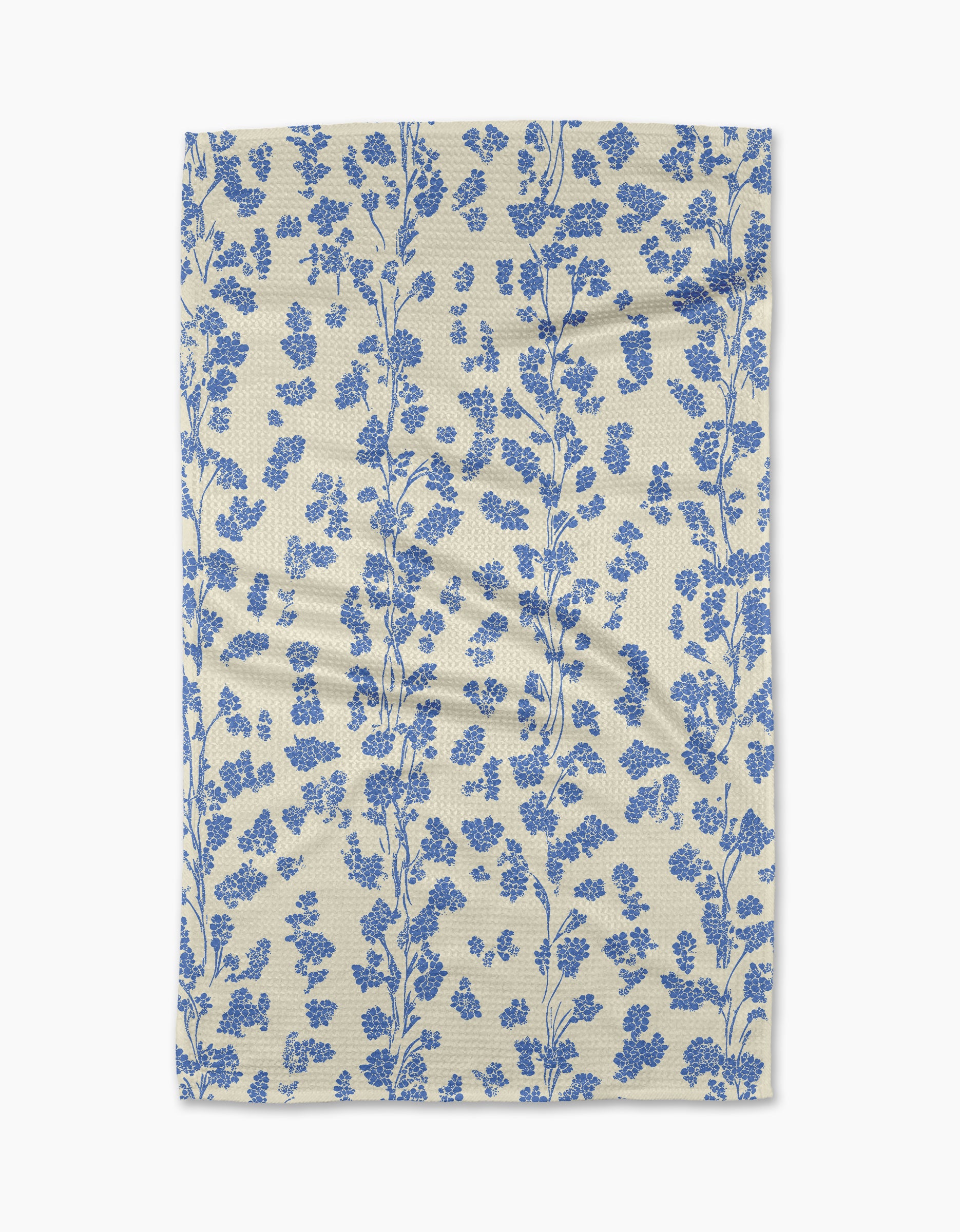 The Lilou Cream kitchen towel by Geometry is eco-friendly and cream-colored, featuring a vertical pattern of blue floral branches with small flowers and leaves. The fabric has a slightly wrinkled look and is shown on a white background.