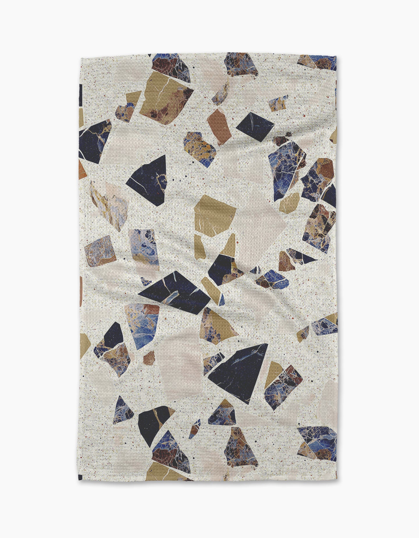 The Geometry Shale blanket features a terrazzo pattern with navy blue, beige, brown, and tan shapes on a speckled off-white background. Its rectangular shape and gently rumpled edges create a cozy, towel-inspired look.