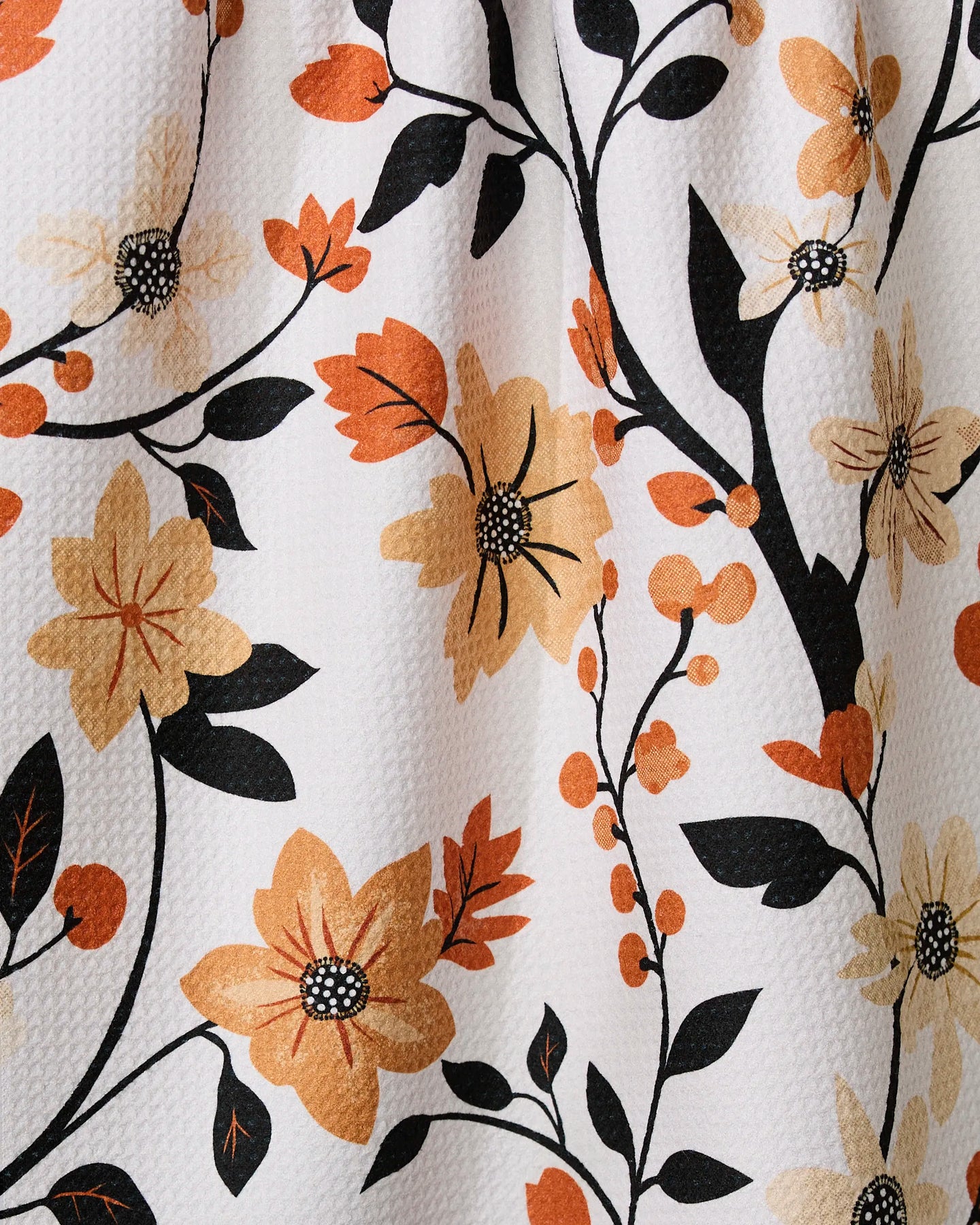 A close-up of the Geometry Spiced Florals tea towel shows its embossed texture and ultra-absorbent design, featuring yellow, orange, and black floral patterns with leaves and stems on a white background.