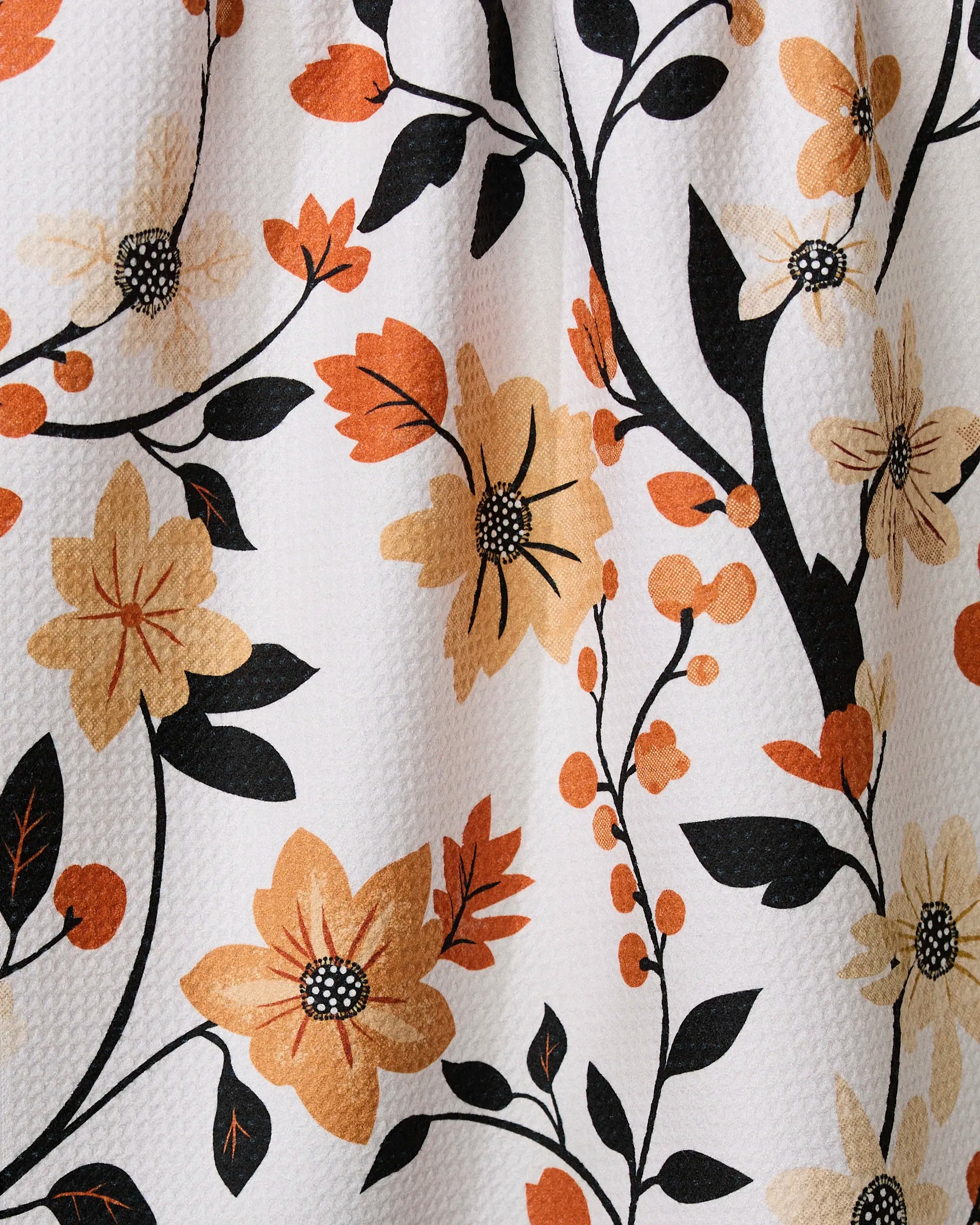 A close-up of the Geometry Spiced Florals tea towel shows its embossed texture and ultra-absorbent design, featuring yellow, orange, and black floral patterns with leaves and stems on a white background.