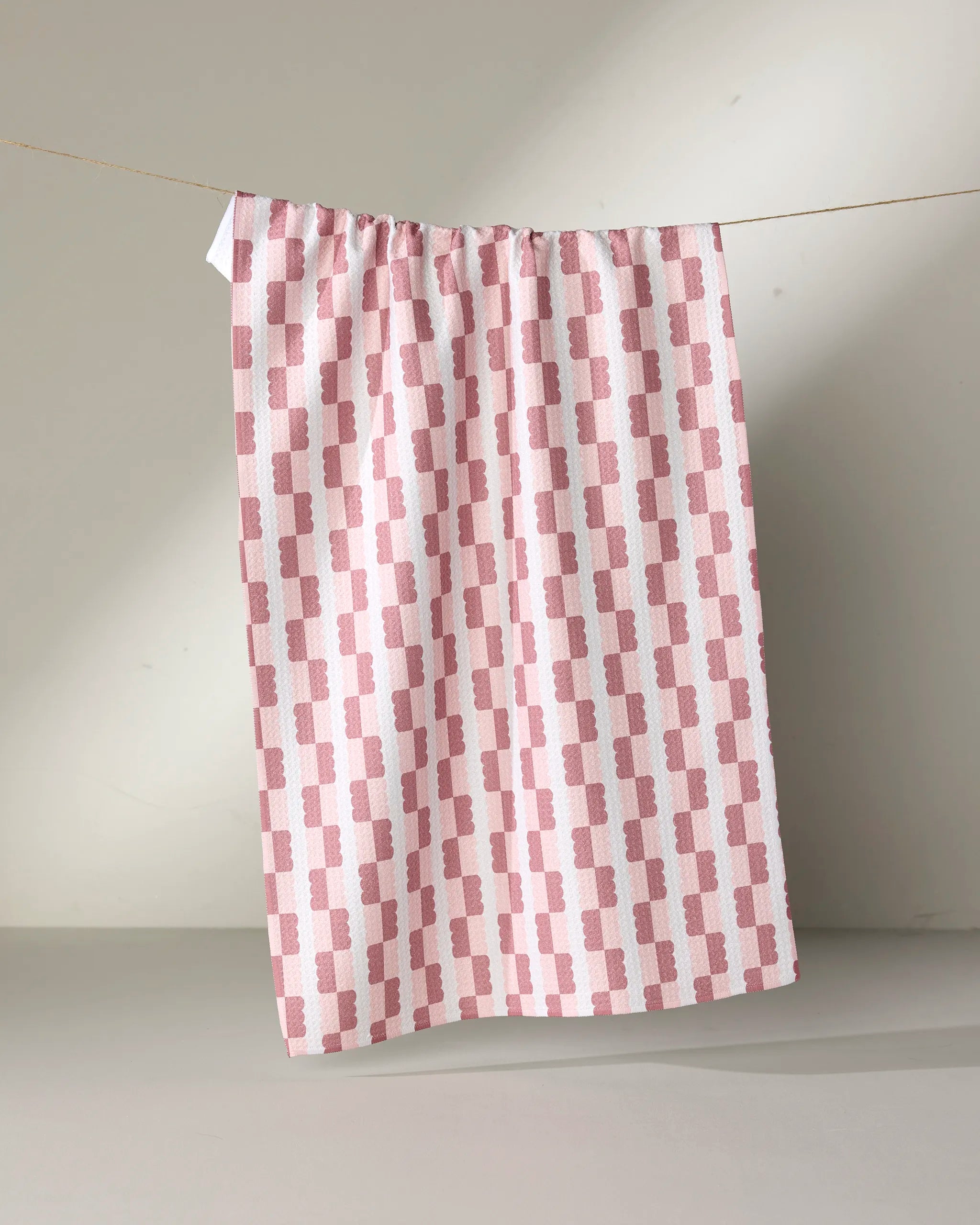 The Brooke Homec "Sweet" kitchen tea towel, featuring a pink and white geometric rectangle pattern, hangs from a string against a plain background. This ultra-absorbent towel combines style and functionality for your kitchen.