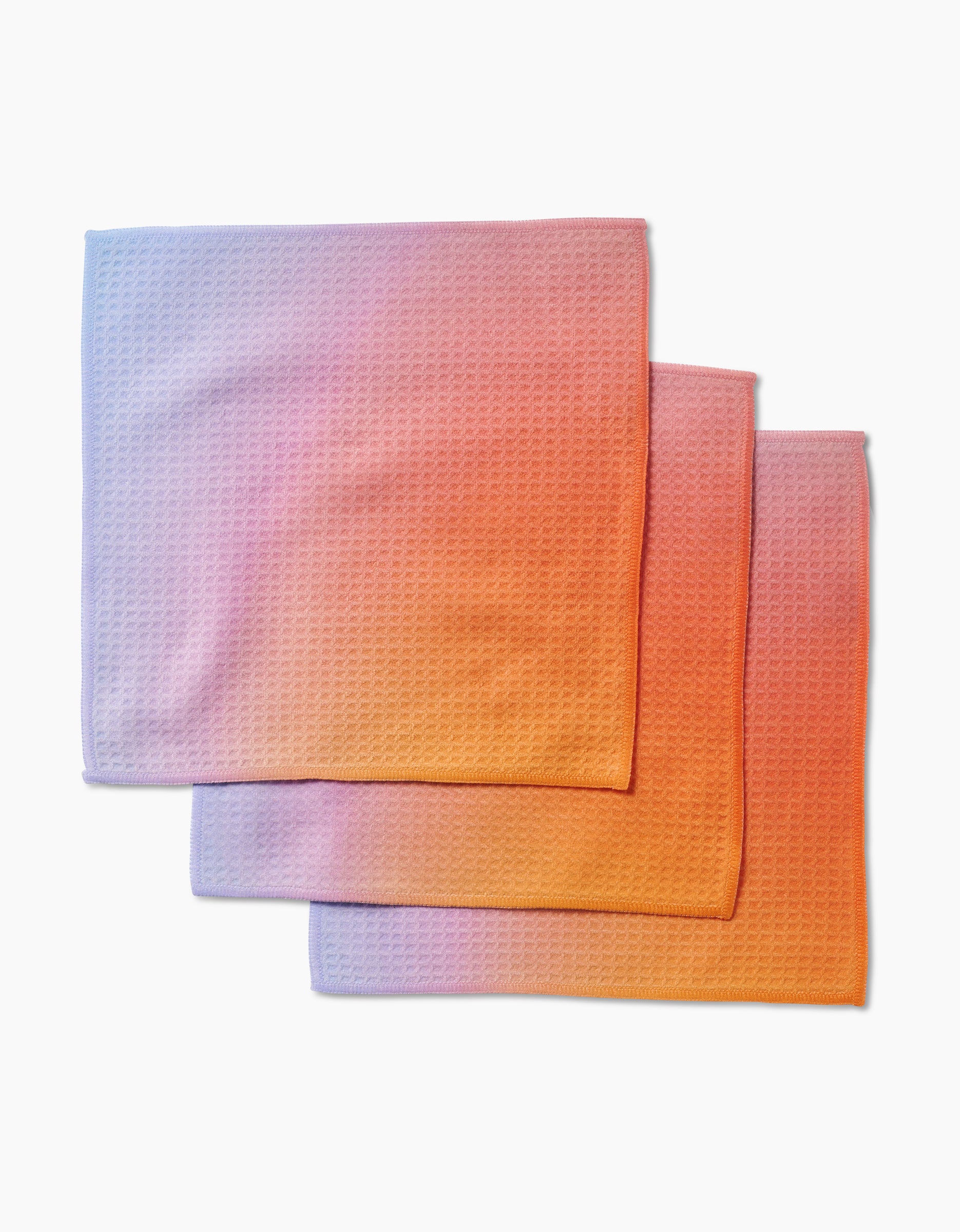Three folded GEOMETRY Aura washcloths, featuring Geoweave technology and a gradient from light purple to orange, are slightly overlapped on a plain white background.